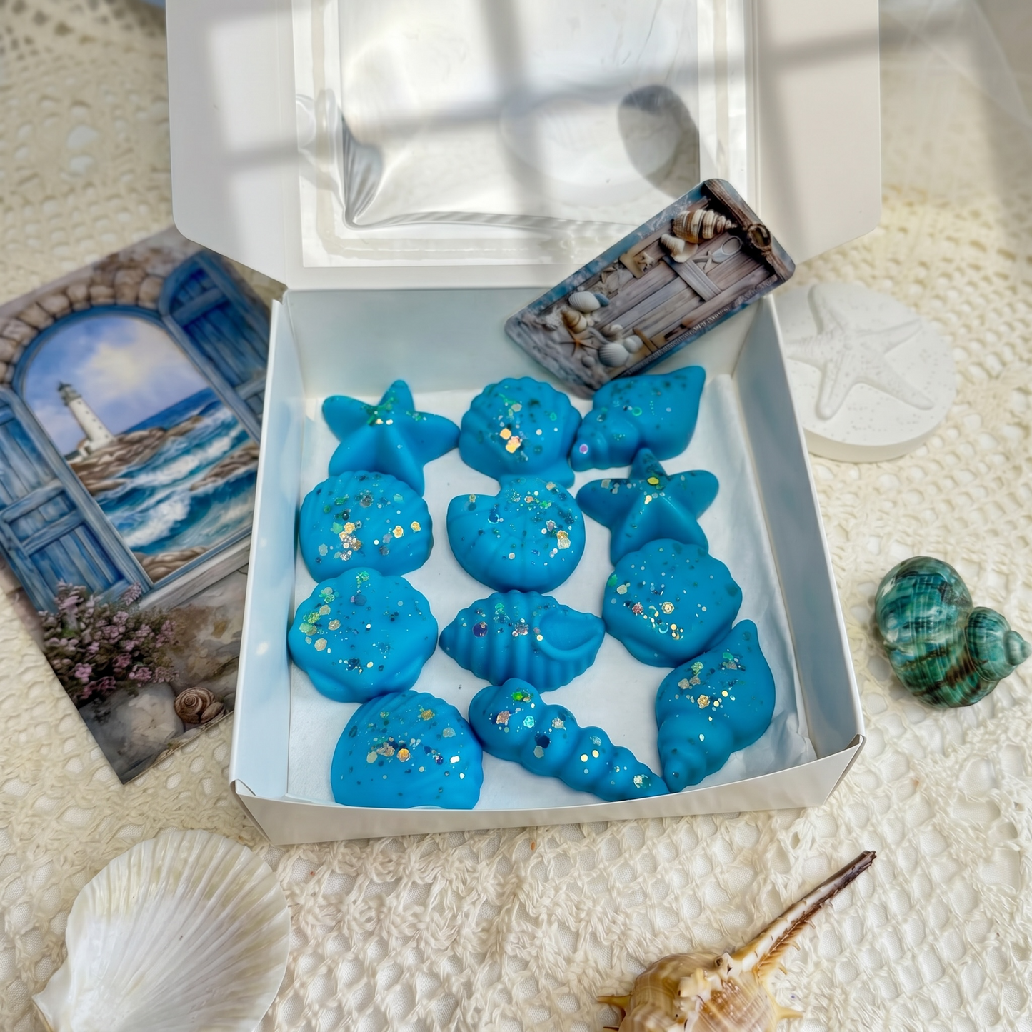 Eine quadratische weiße Geschenkbox mit geöffnetem Sichtfenster, gefüllt mit elf hellblauen Wachsschmelzen (Wax Melts) mit goldenem Glitzer in maritimen Formen, umgeben von einer Postkarte mit Leuchtturmmotiv, echten Seemuscheln und einem weißen Deko-Seestern.