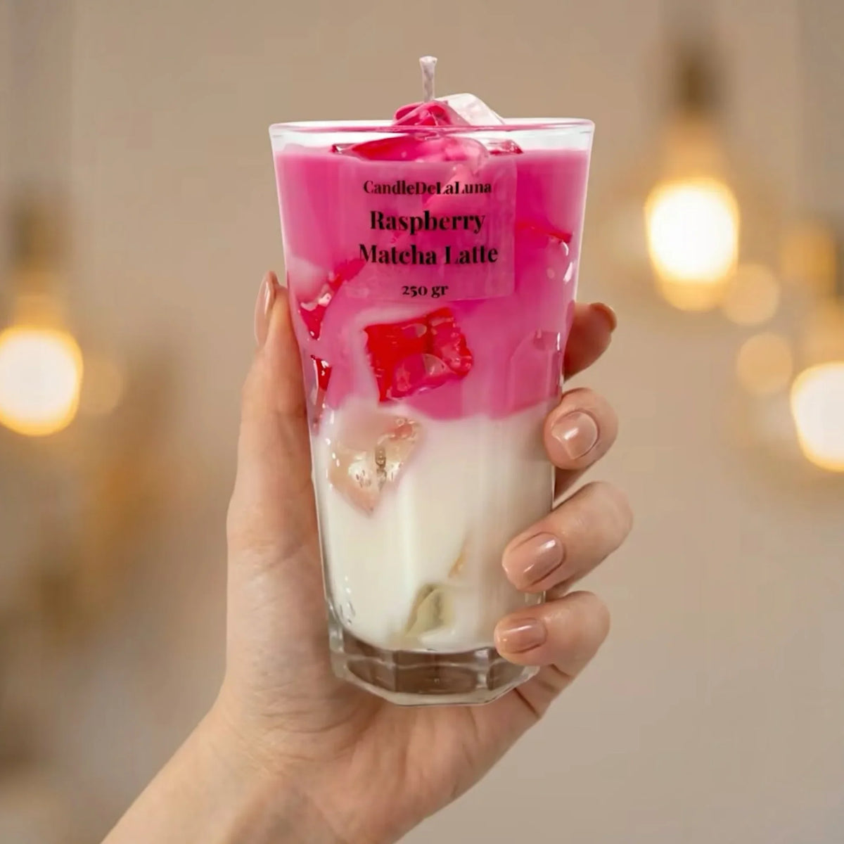 Eine Hand hält eine dekorative Kerze in einem hohen, facettierten Glas, die optisch einem geschichteten Raspberry Matcha Latte mit einer intensiven rosa oberen Schicht, einer weißen unteren Schicht und Wachs-Eiswürfeln ähnelt.