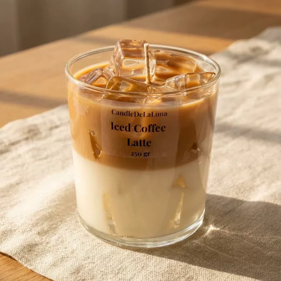 Eine dekorative Kerze in Form eines Iced Coffee Latte in einem niedrigen Glas steht auf einer hellen Tischdecke aus Leinen, beleuchtet von warmem, natürlichem Sonnenlicht.