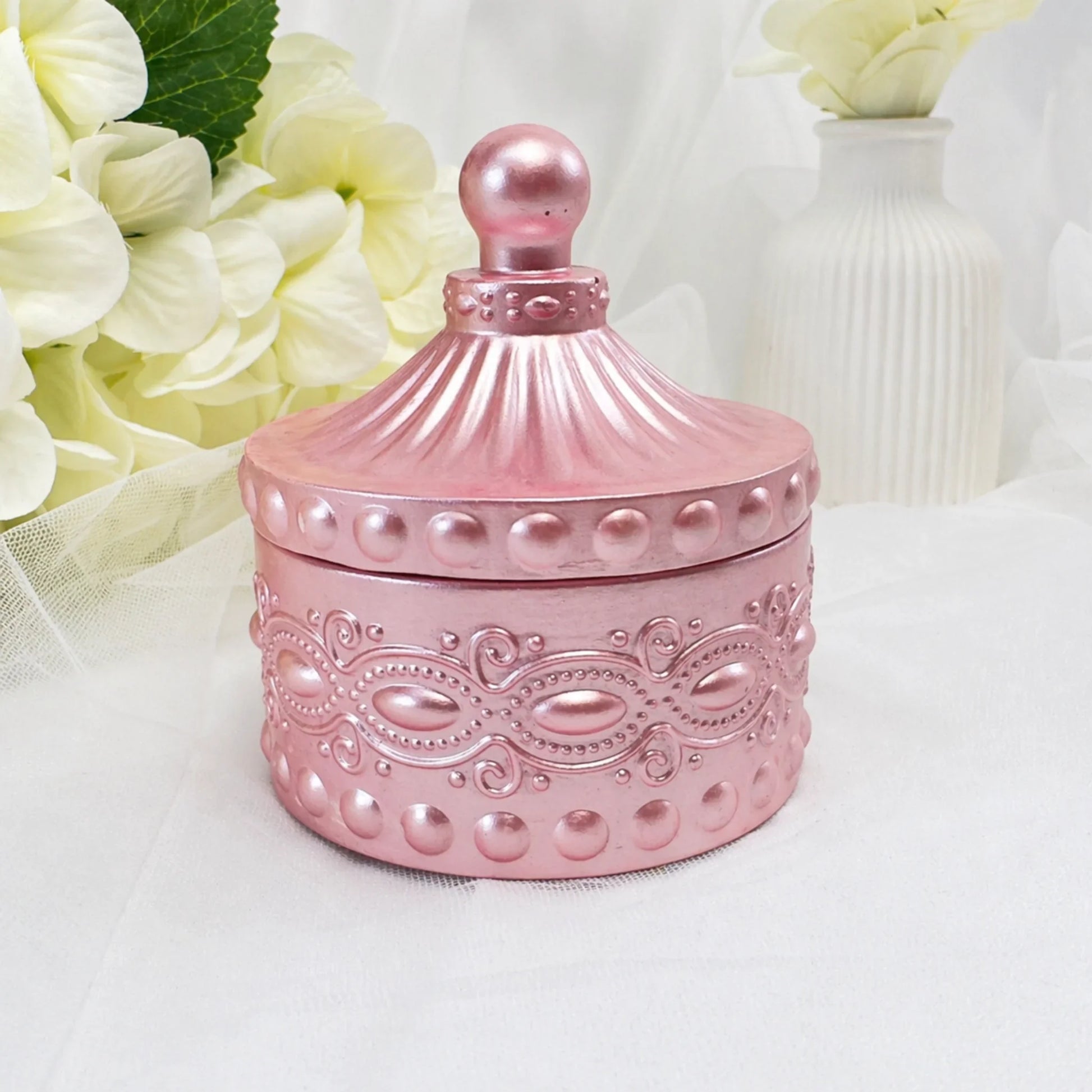 Eine geschlossene Duftkerze in einem eleganten, rosa-metallicfarbenen Gefäß mit aufwendigen Ornamenten und einem passenden spitzen Deckel, präsentiert vor einem hellen Hintergrund mit weißen Hortensien.
