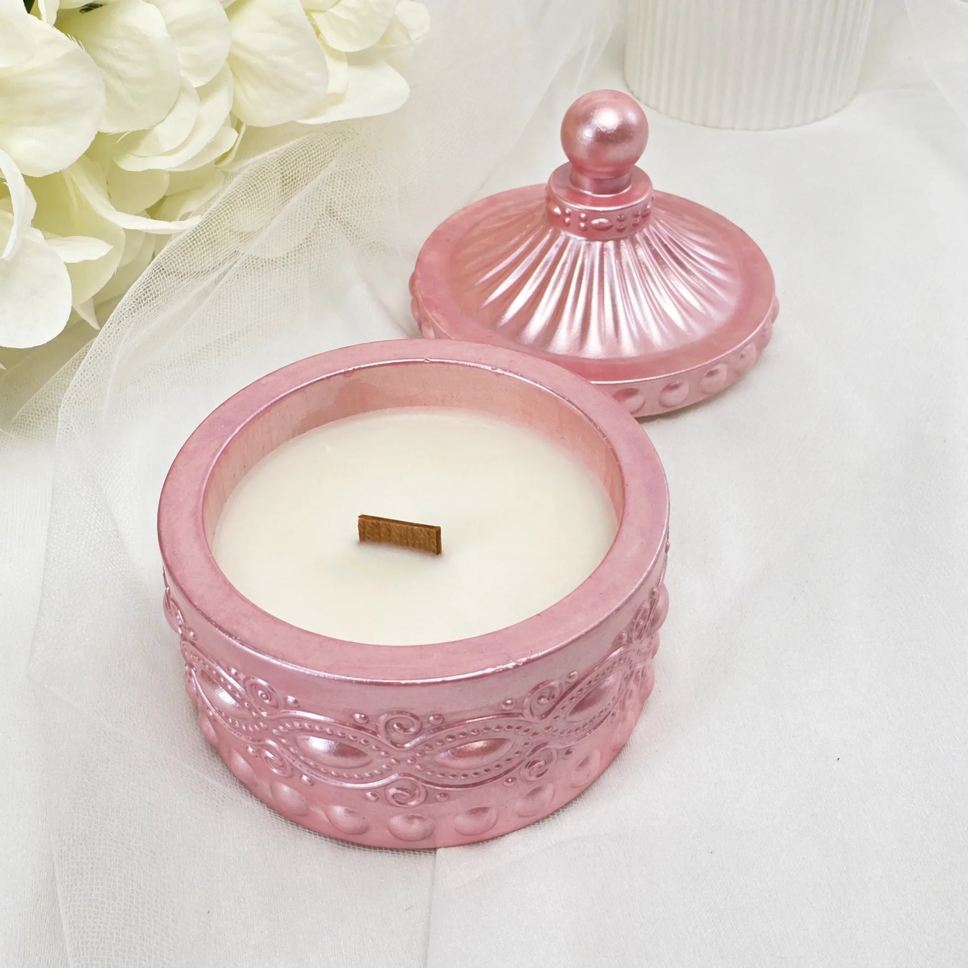 Eine geöffnete, rosa-metallicfarbene Duftkerze in einem verzierten Glasgefäß mit passendem Deckel, weißem Wachs und einem Holzdocht, platziert auf einem hellen Stoffhintergrund neben weißen Blumen.