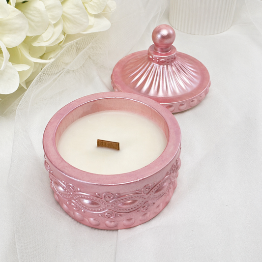 Eine geöffnete, rosa-metallicfarbene Duftkerze in einem verzierten Glasgefäß mit passendem Deckel, weißem Wachs und einem Holzdocht, platziert auf einem hellen Stoffhintergrund neben weißen Blumen.