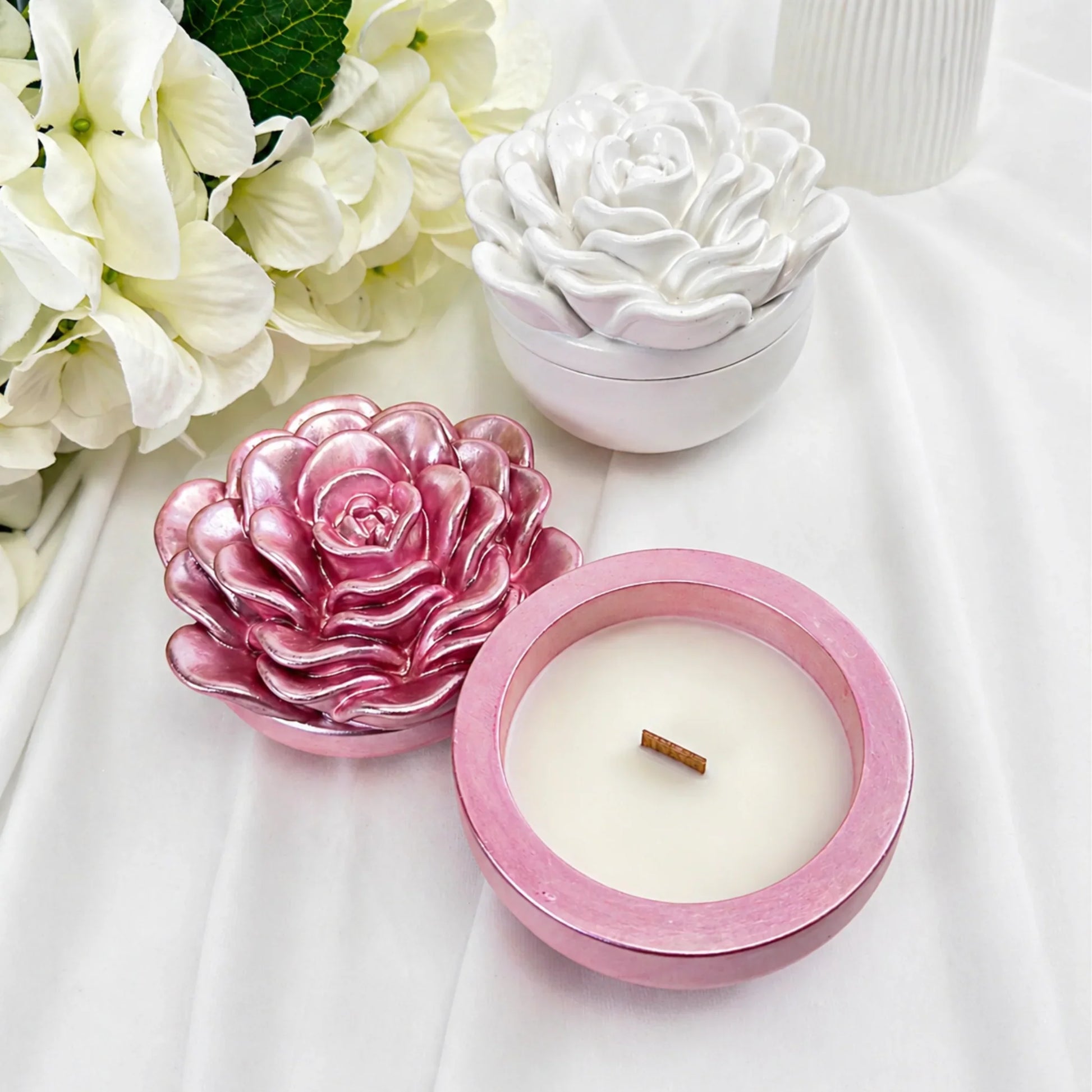 Detailansicht einer geöffneten rosa Keramik-Duftkerze mit Holzdocht und einer geschlossenen weißen Kerze mit Kameliendeckel auf hellem Stoff.
