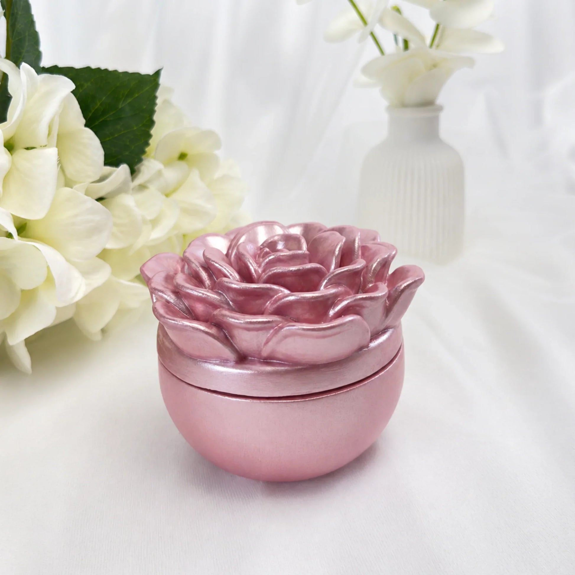Elegante rosa Duftkerze in einer runden Keramikschale mit kunstvollem Deckel in Kamelienform, dekoriert mit weißen Blumen im Hintergrund.