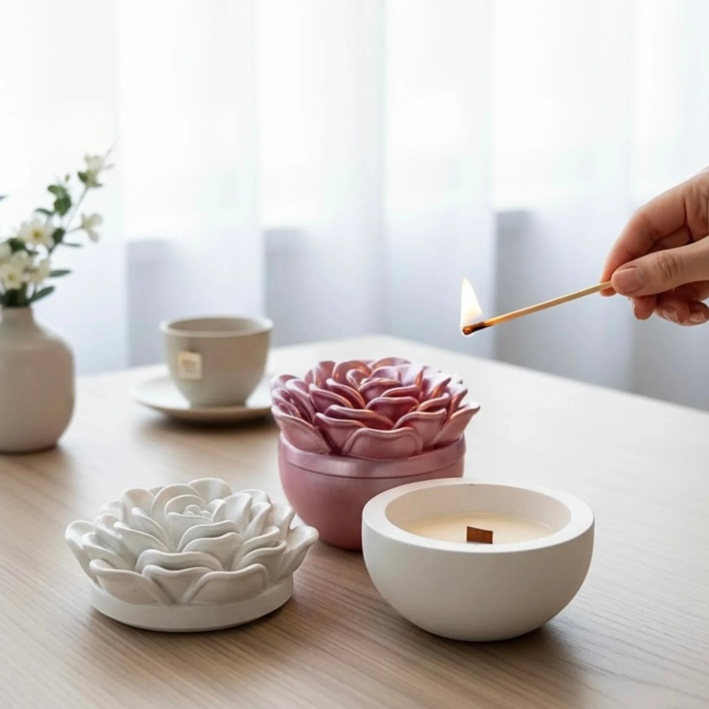 Gemütliche Atmosphäre mit einer angezündeten Sojakerze im weißen Keramiktopf, daneben eine geschlossene rosa Kamelien-Kerze auf einem Holztisch mit Teetasse.