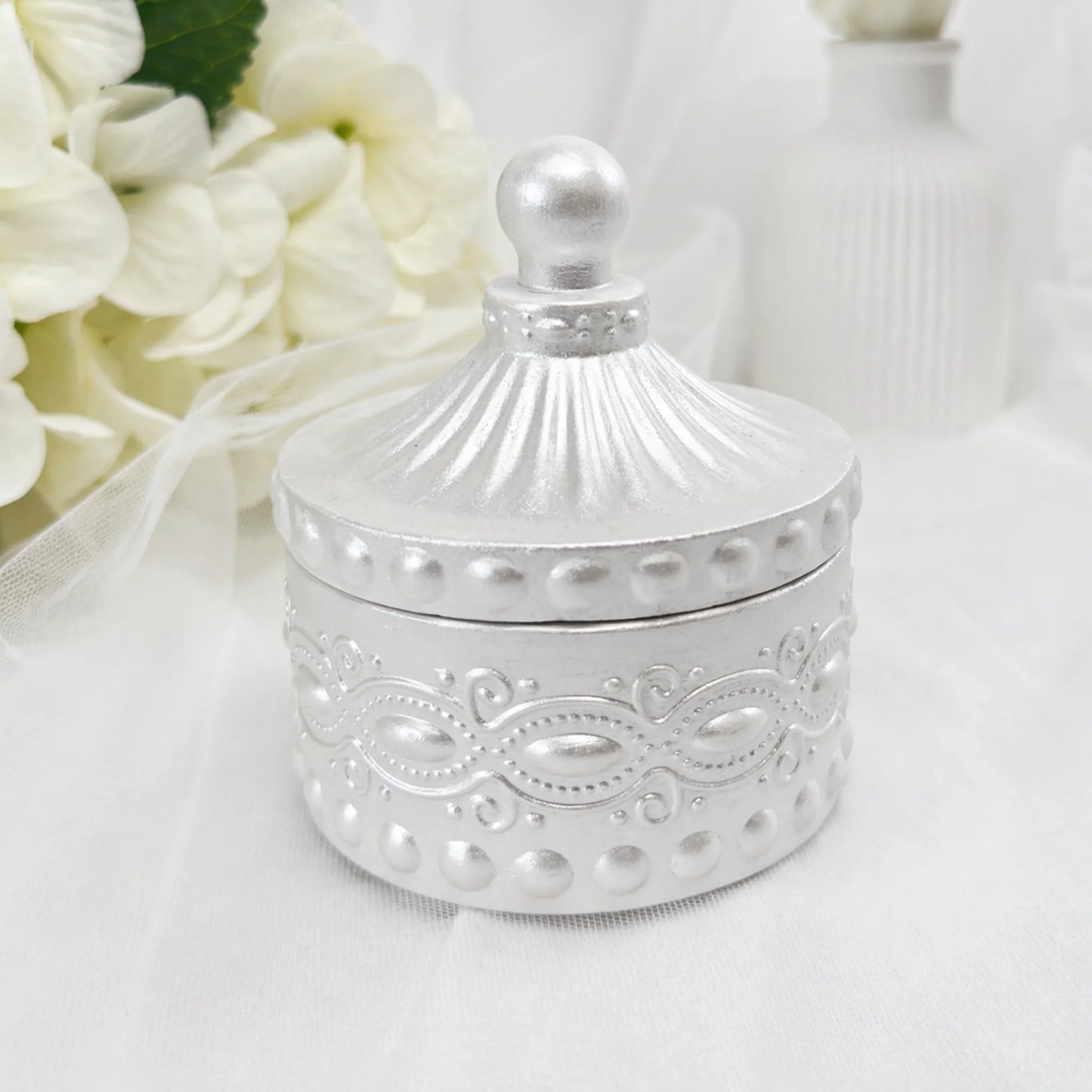Eine geschlossene, silberne Duftkerze in einem reich verzierten Gefäß mit passendem Deckel, elegant präsentiert vor einem Hintergrund aus weißen Hortensien und einer weißen Vase.