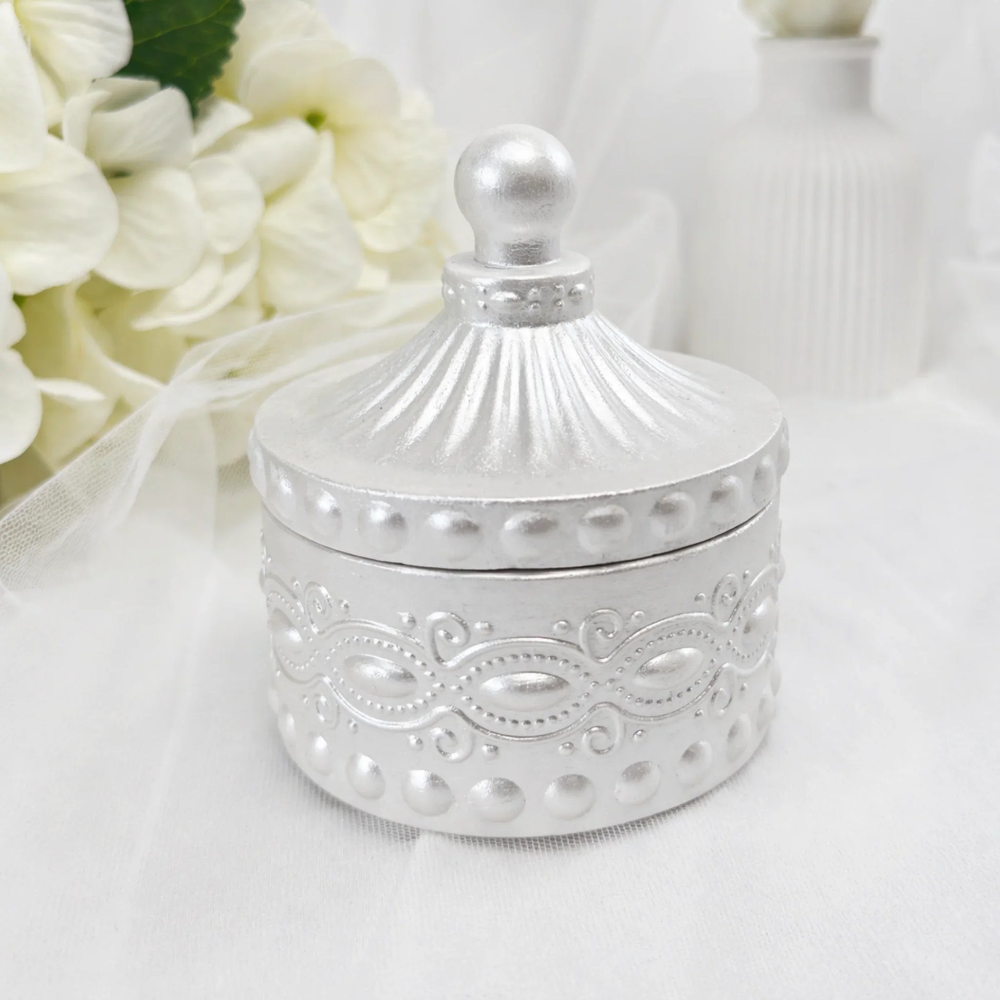 Eine geschlossene, silberne Duftkerze in einem reich verzierten Gefäß mit passendem Deckel, elegant präsentiert vor einem Hintergrund aus weißen Hortensien und einer weißen Vase.