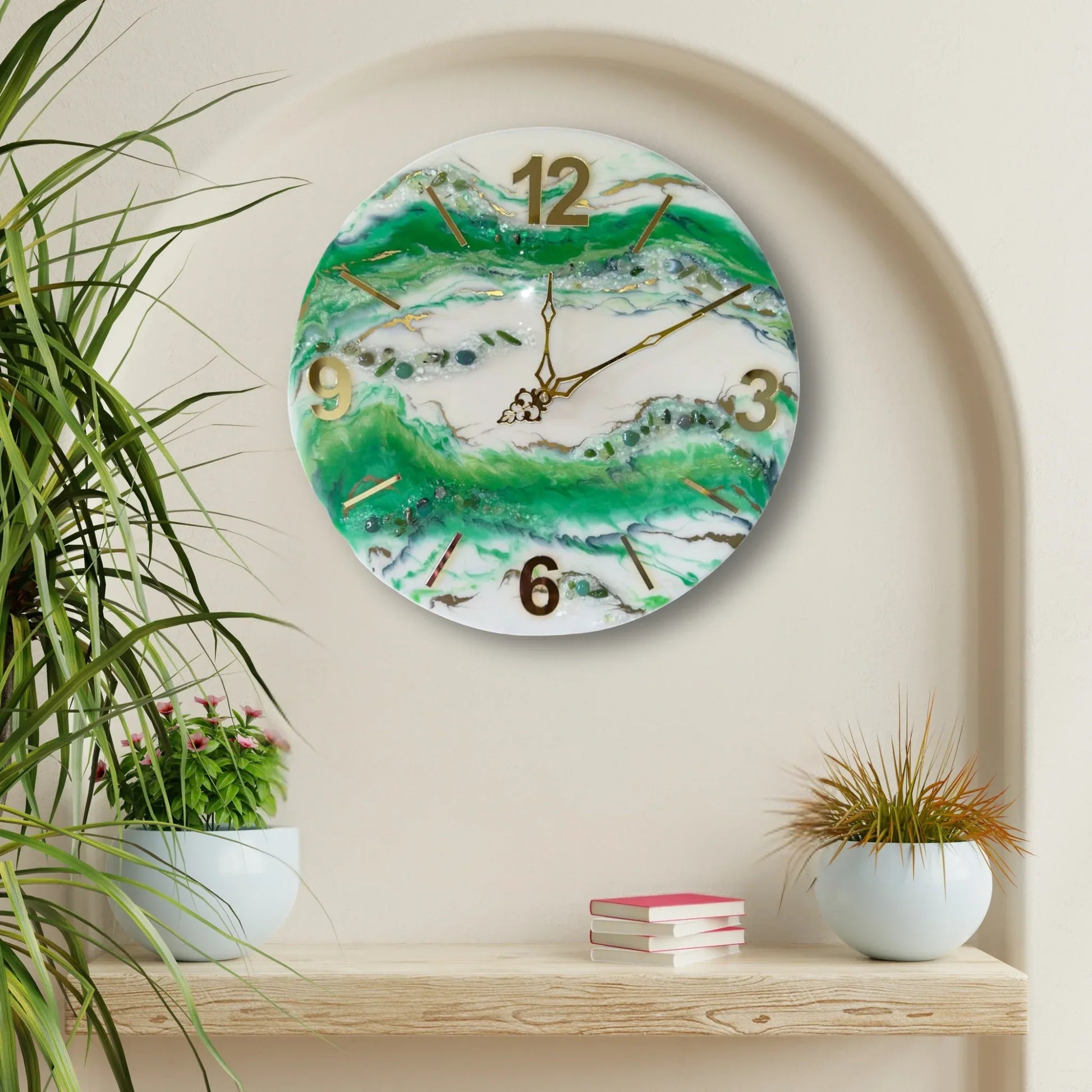 Eine runde Wanduhr aus Epoxidharz im Geoden-Stil mit grünen, weißen und goldenen Mustern, aufgehängt in einer ovalen Wandnische über einem Holzregal mit Büchern und zwei Topfpflanzen in hellen Keramiktöpfen.