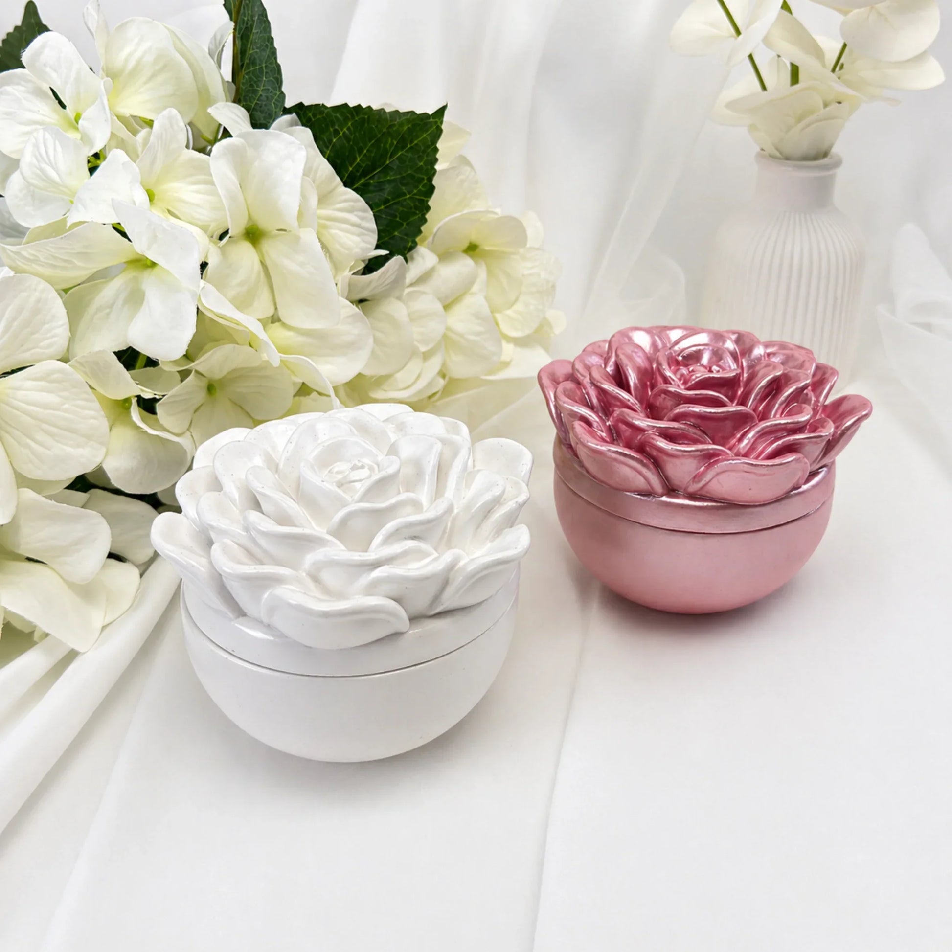 Zwei handgemachte Duftkerzen in Weiß und schimmerndem Rosa mit dekorativen Kameliendeckeln neben einem weißen Blumenstrauß auf hellem Untergrund.
