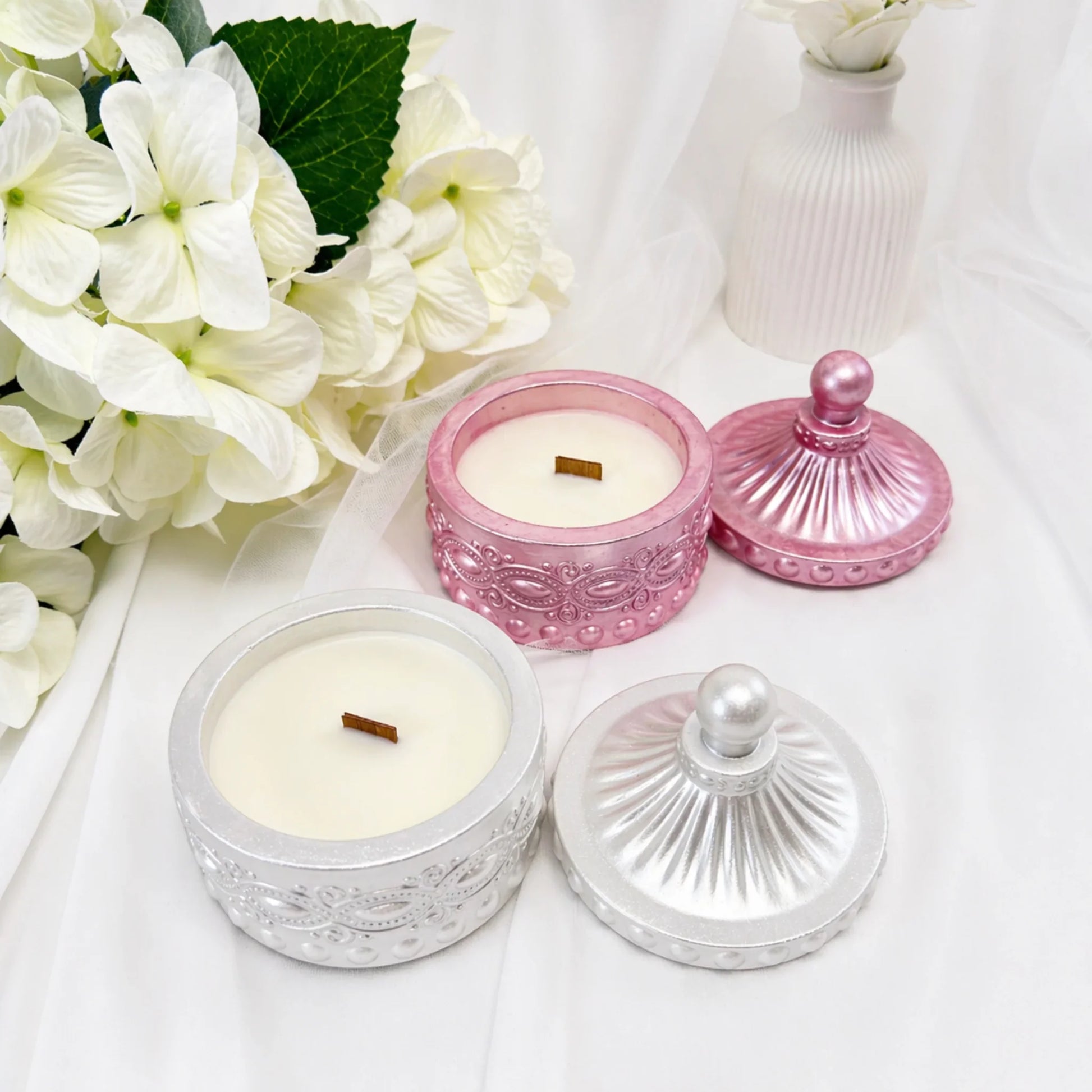 Zwei geöffnete Duftkerzen in Silber und Rosa-Metallic mit sichtbaren Holzdochten und danebenliegenden Deckeln, arrangiert mit weißen Hortensien und einer dekorativen Vase auf hellem Stoff.
