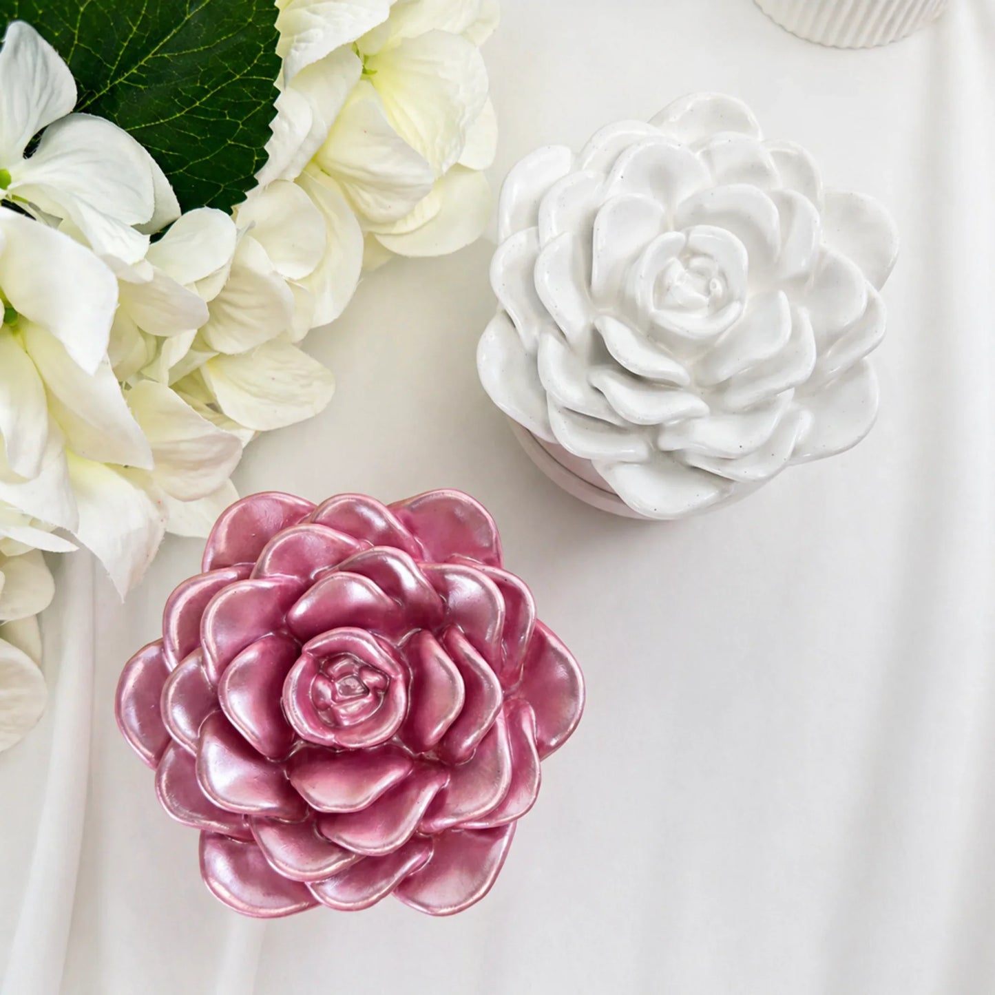 Draufsicht auf zwei geschlossene Keramik-Duftkerzen in Rosa und Weiß mit kunstvollem Deckel in Kamelien-Design auf weißem Hintergrund.