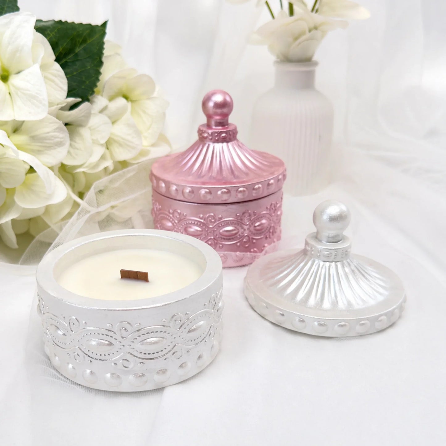 Zwei stilvolle Duftkerzen in verzierten Gefäßen – eine geöffnete silberne Kerze mit Holzdocht im Vordergrund und eine geschlossene rosa-metallic Kerze dahinter, dekoriert mit weißen Hortensien und einer weißen Vase.
