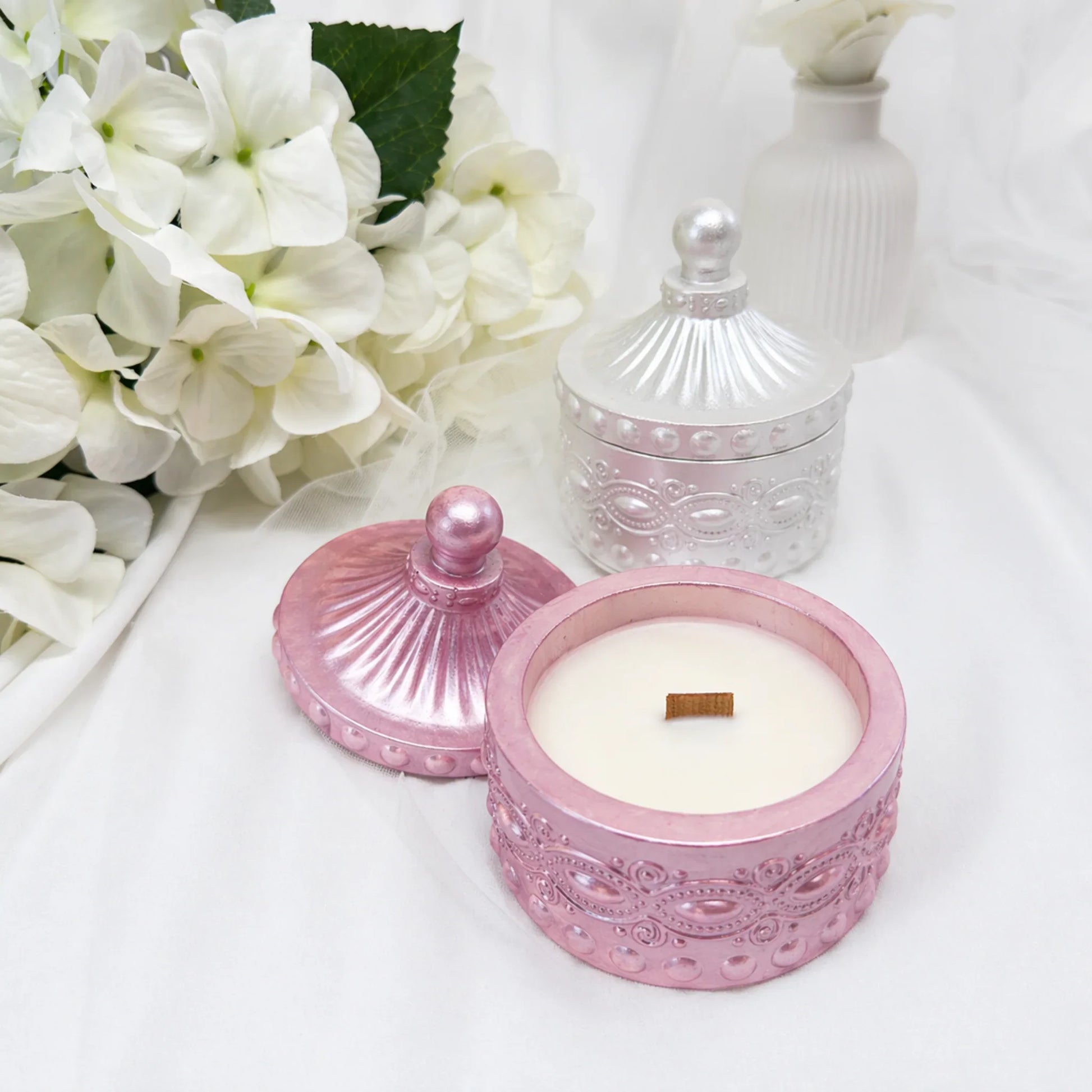 Zwei elegante Duftkerzen in verzierten Gefäßen – eine geöffnete rosa-metallic Kerze mit Holzdocht im Vordergrund und eine geschlossene silberne Kerze dahinter, dekoriert mit weißen Hortensien.
