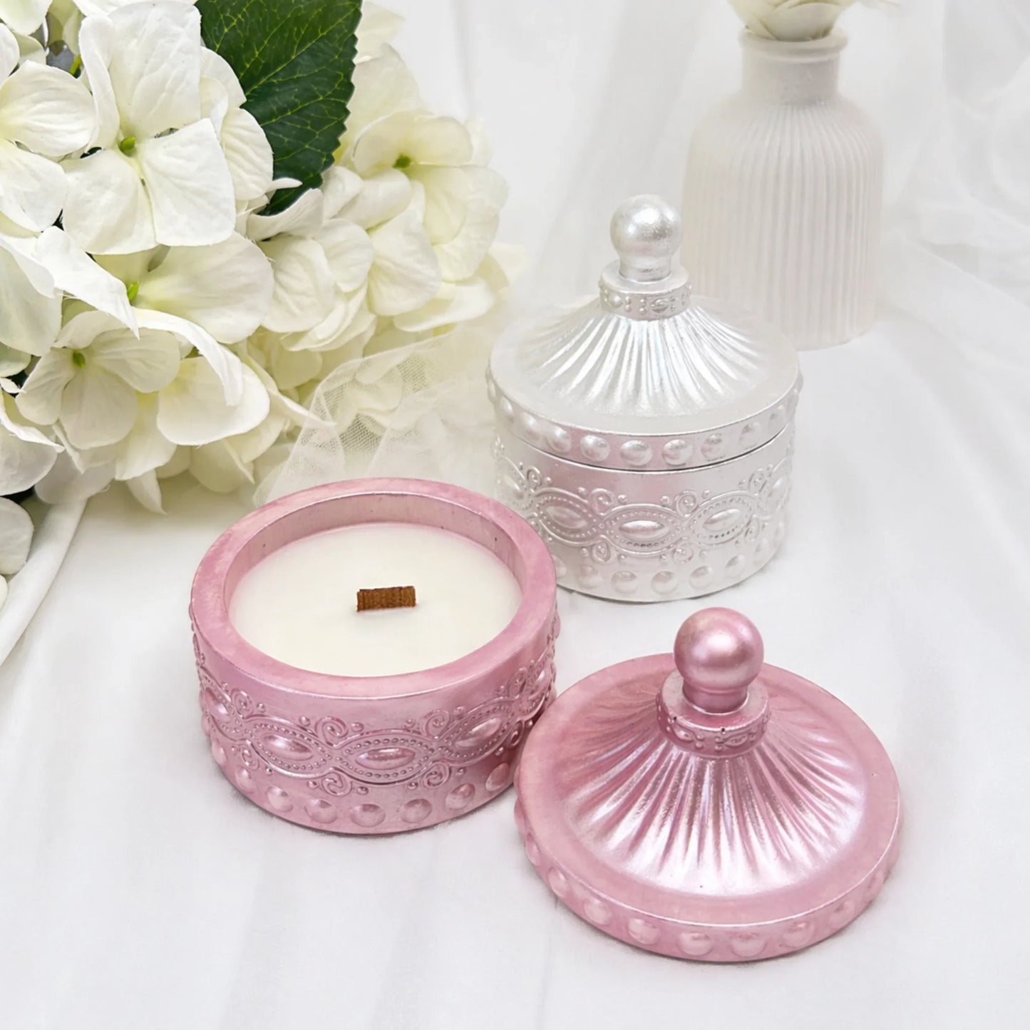 Zwei dekorative Duftkerzen in rosa-metallic und perlmutt-weiß auf hellem Hintergrund, wobei die rosa Kerze im Vordergrund geöffnet ist und ihren Holzdocht zeigt, umrahmt von weißen Blumen.
