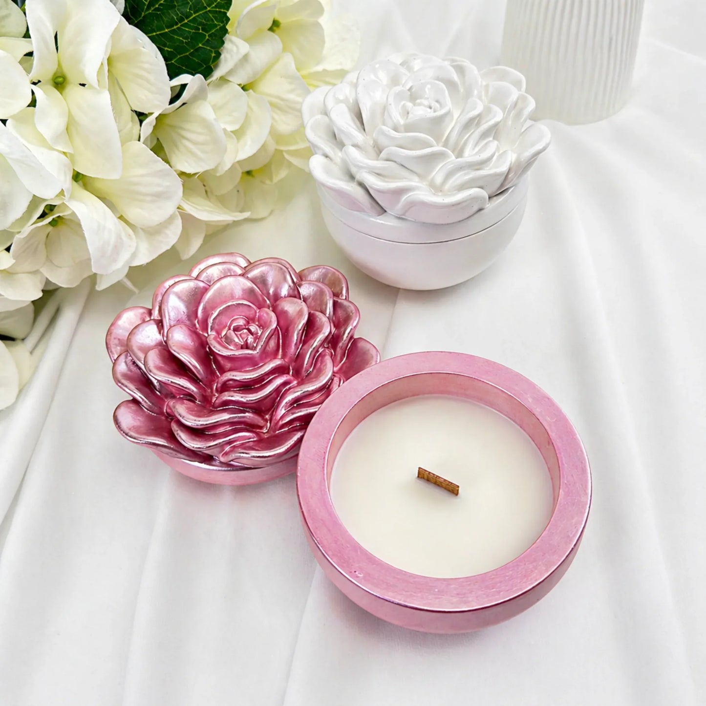 Nahaufnahme einer rosa Sojawachskerze mit sichtbarem Holzdocht und schimmerndem Kameliendeckel neben einer weißen Keramikdose auf weißem Seidenstoff.