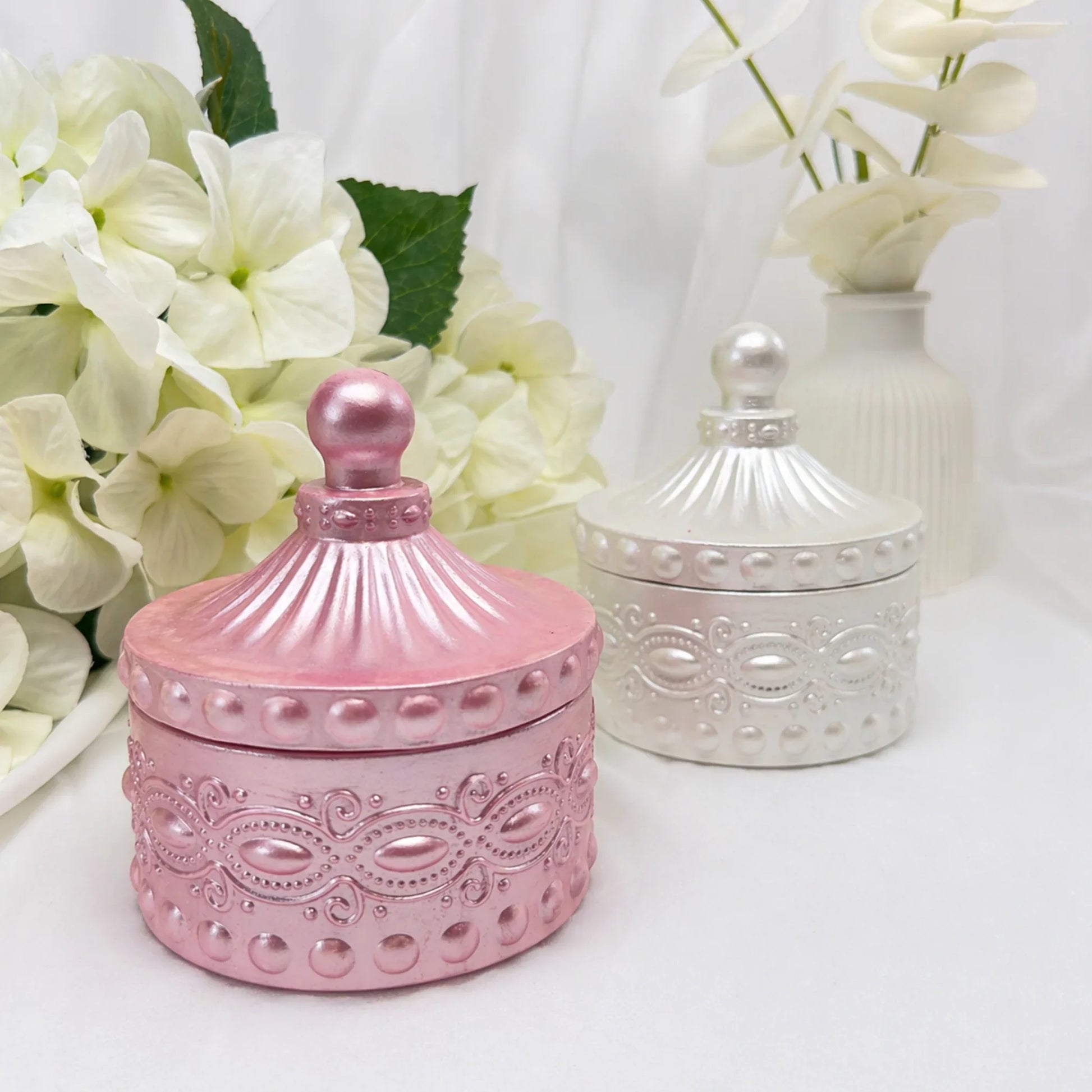 Zwei geschlossene Duftkerzen in eleganten Gefäßen – eine im Vordergrund in Rosa-Metallic und eine im Hintergrund in Perlmutt-Silber, stilvoll arrangiert mit weißen Hortensien und einer weißen Vase.