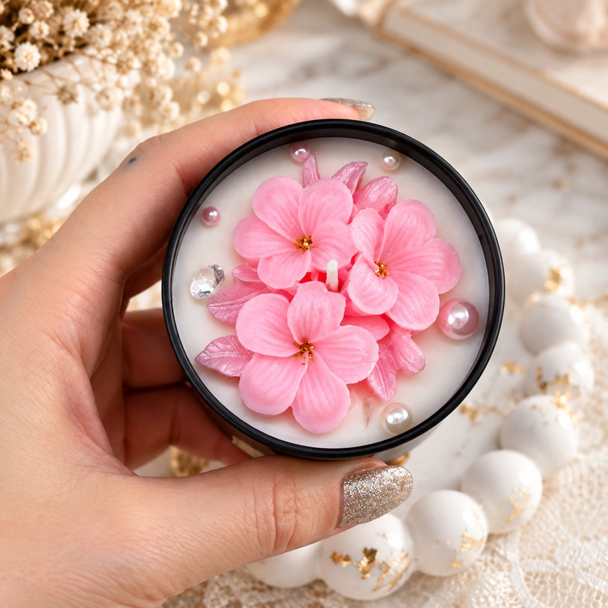 Kerze in schwarzer Dose mit rosa Blüten und kleinen Perlen auf weißer Oberfläche
