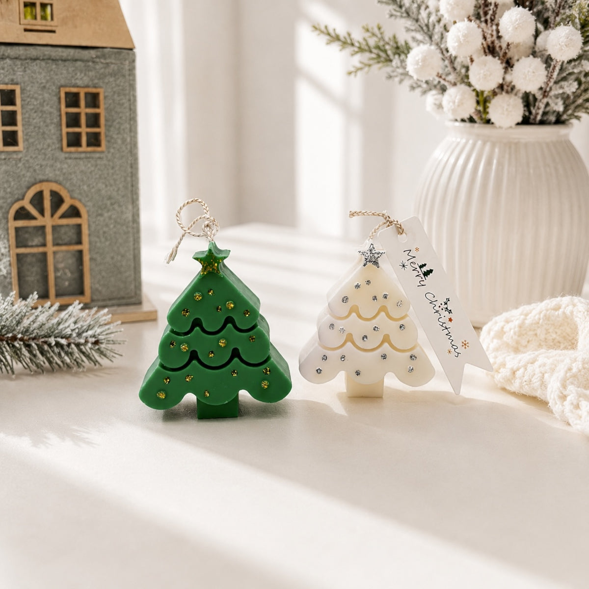 Grüner und weißer Weihnachtsbaum-Anhänger mit Sternspitzen, glitzernden Punkten und Schlaufen vor einem Dekohäuschen
