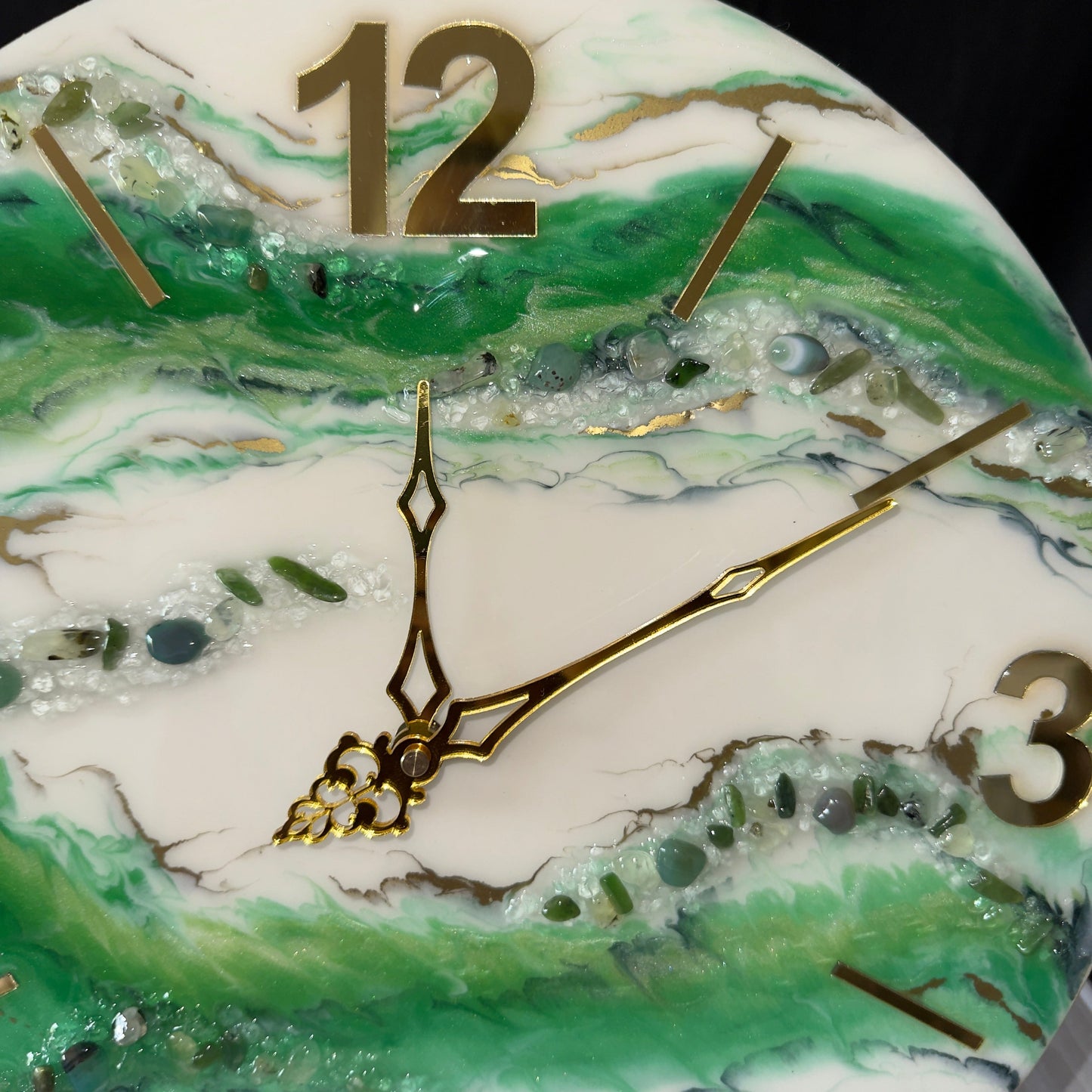 Nahaufnahme einer Wanduhr aus Epoxidharz im Geoden-Stil mit grünen und weißen Mustern, goldenen Zeigern, einer großen goldenen Ziffer 12 im oberen Bereich sowie eingebetteten grünen Dekosteinen, Glitzer und goldenen Adern auf der Oberfläche.