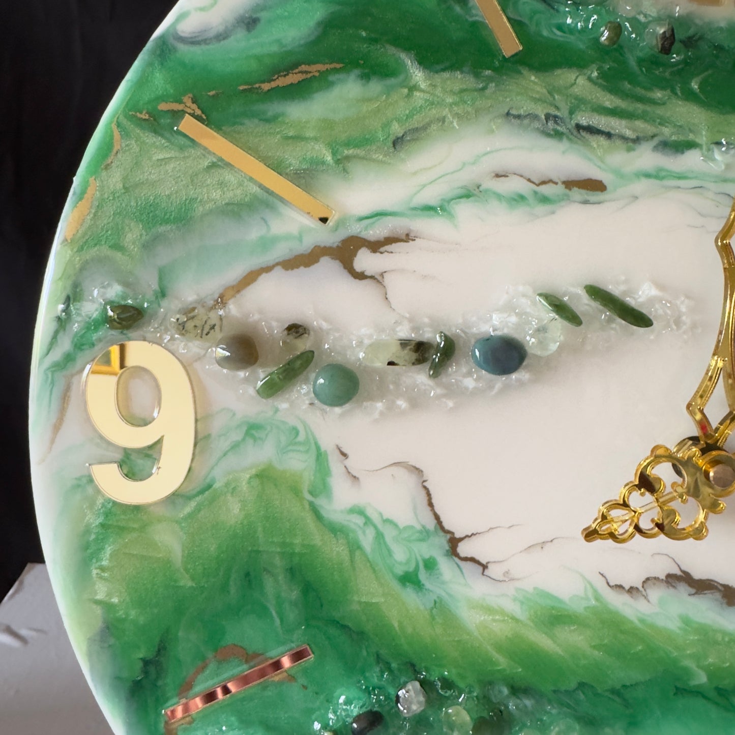 Nahaufnahme einer Wanduhr aus Epoxidharz im Geoden-Stil mit grünen und weißen Mustern, einer großen goldenen Ziffer 9 auf der linken Seite, goldenen Zeigern sowie einer Linie aus eingebetteten grünen Dekosteinen, Glitzer und goldenen Linien.