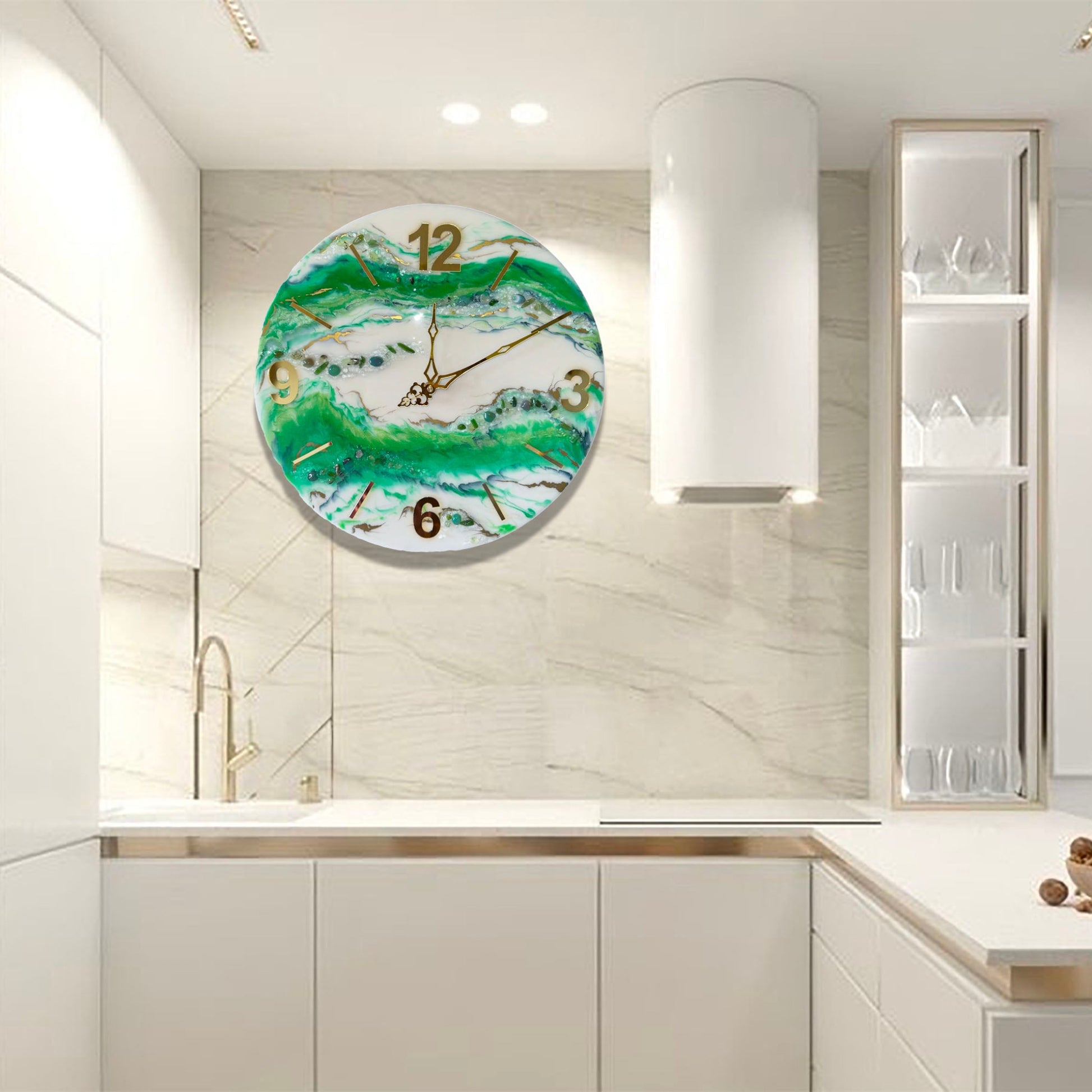 Eine runde Wanduhr aus Epoxidharz im Geoden-Stil mit grünen, weißen und goldenen Mustern, aufgehängt an einer hellen Marmorwand in einer modernen weißen Küche neben einer zylindrischen Dunstabzugshaube und einem offenen Regal.