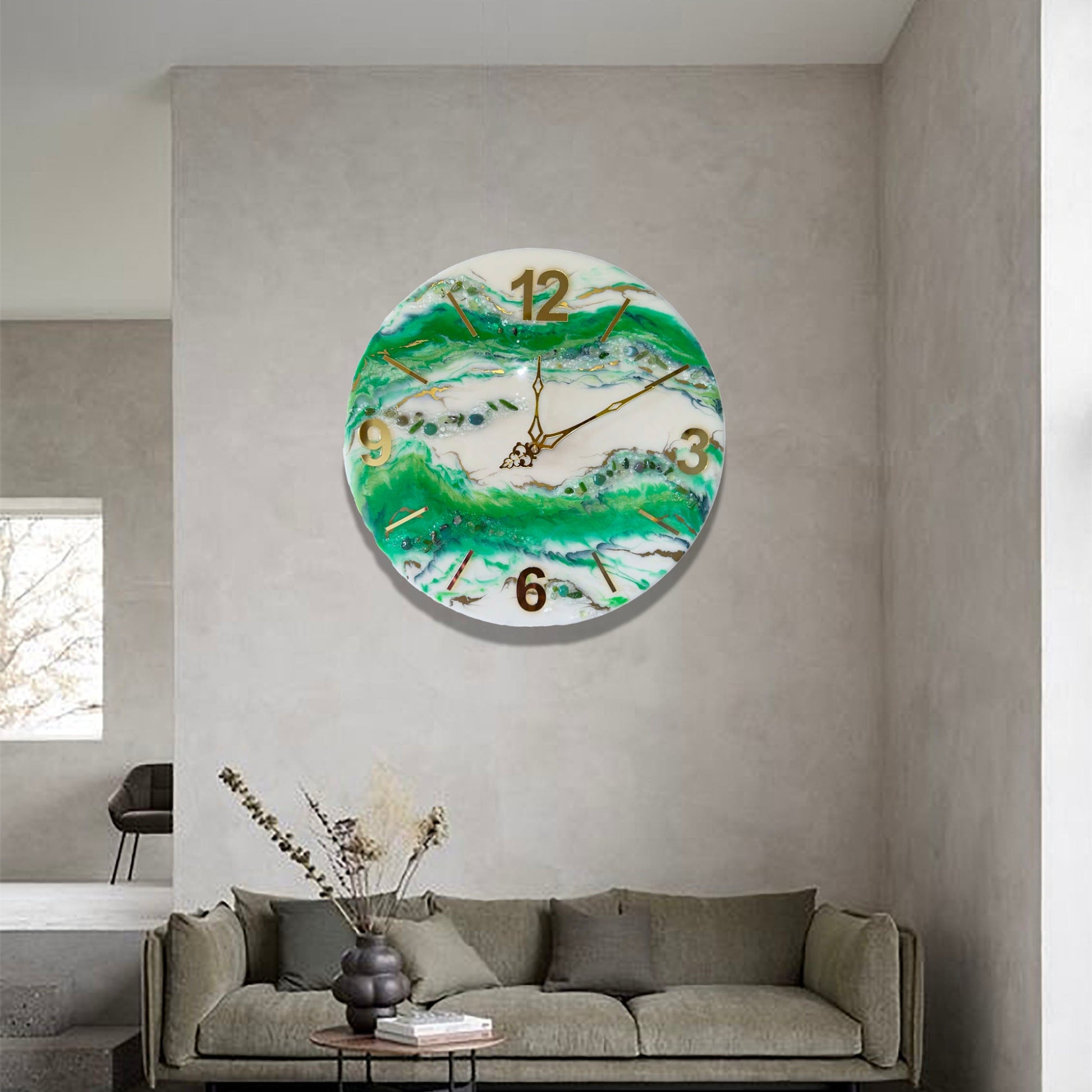 Eine runde Wanduhr aus Epoxidharz im Geoden-Stil mit grünen, weißen und goldenen Mustern, aufgehängt an einer minimalistischen grauen Betonwand über einem modernen Sofa mit Kissen und einem kleinen runden Dekotisch.