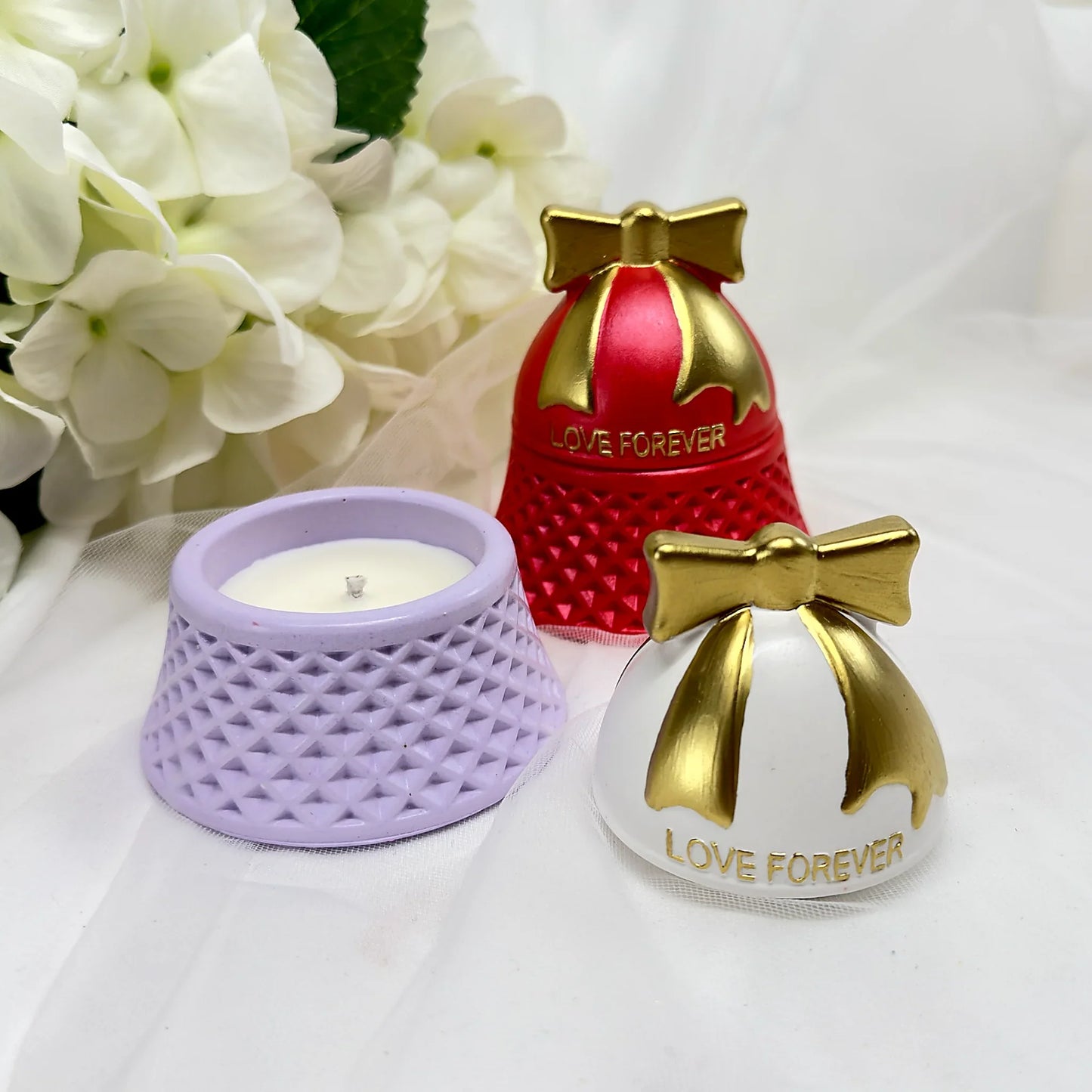 Nahaufnahme von „Love Forever“ Kerzen in Rot und Lavendel mit goldenen Schleifen-Deckeln, eine Kerze geöffnet auf weißem Tüllhintergrund.