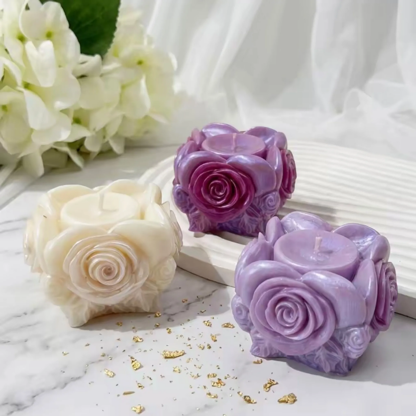 Drei Rosenkerzen in Weiß, Lila und Violett auf Marmortisch mit Hortensien und Goldfolie
