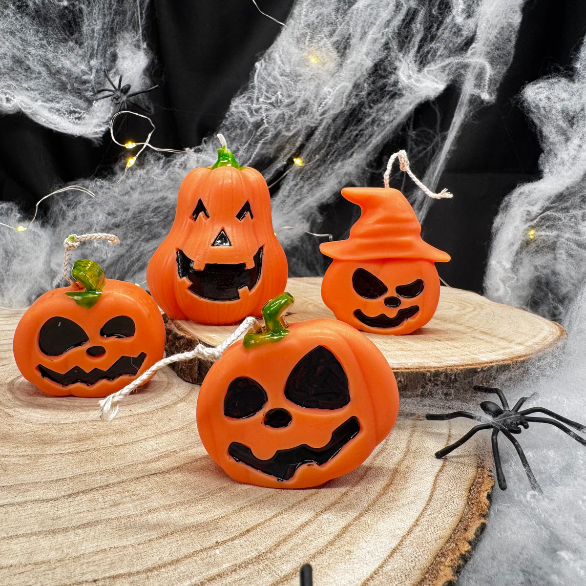 Vier Jack O’Lantern Kürbiskerzen, eine mit orangem Hexenhut, auf Holzscheibe vor Spinnweben und Lichterkette
