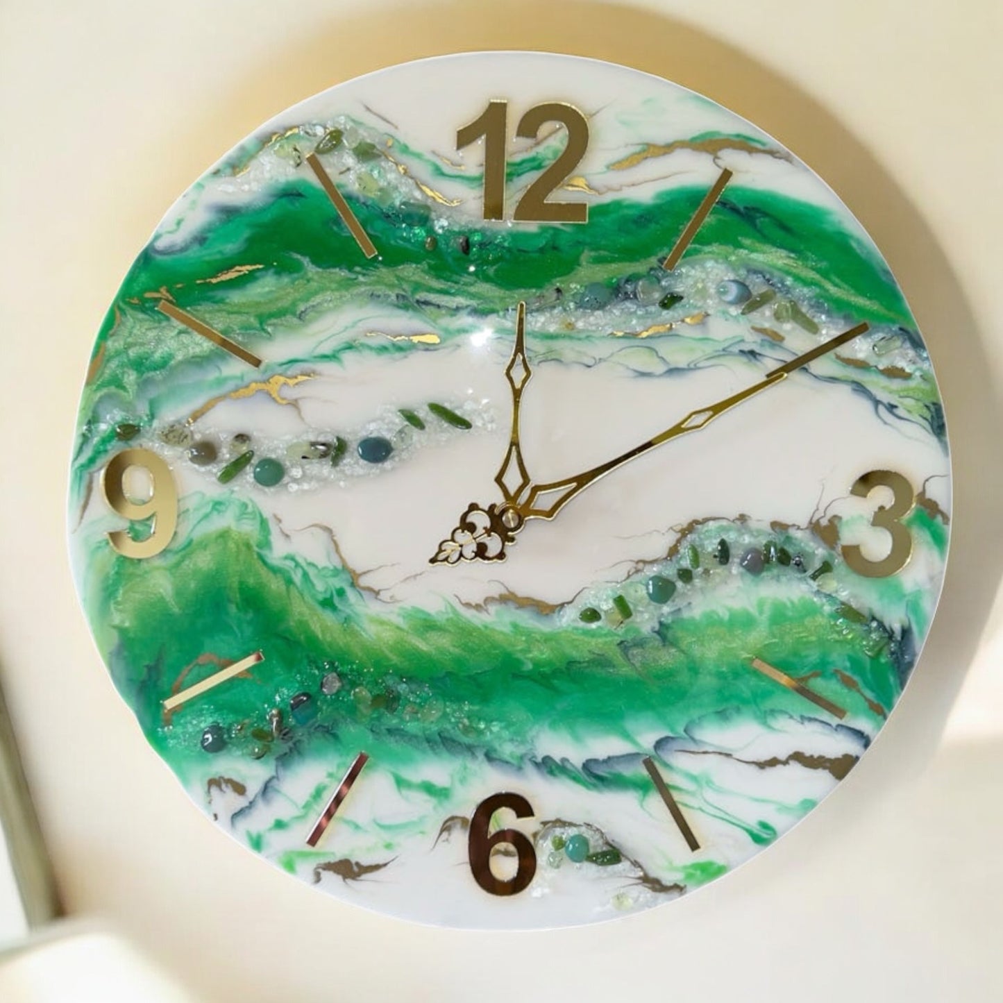 Vorderansicht einer runden Wanduhr aus Epoxidharz im Geoden-Stil mit grünen, weißen und goldenen Mustern, goldenen Zeigern, erhabenen Ziffern (12, 3, 6, 9) und Strichindizes sowie eingebetteten Dekosteinen auf hellem Hintergrund.