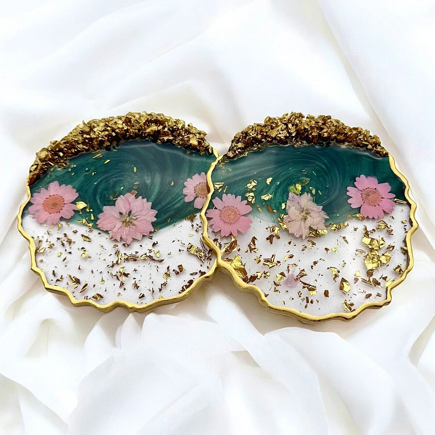 Zwei handgemachte Resin Untersetzer mit rosa Blüten, Blattgold-Akzenten und einem luxuriösen Rand aus goldfarbenen Kristallen auf weißem Stoff.
