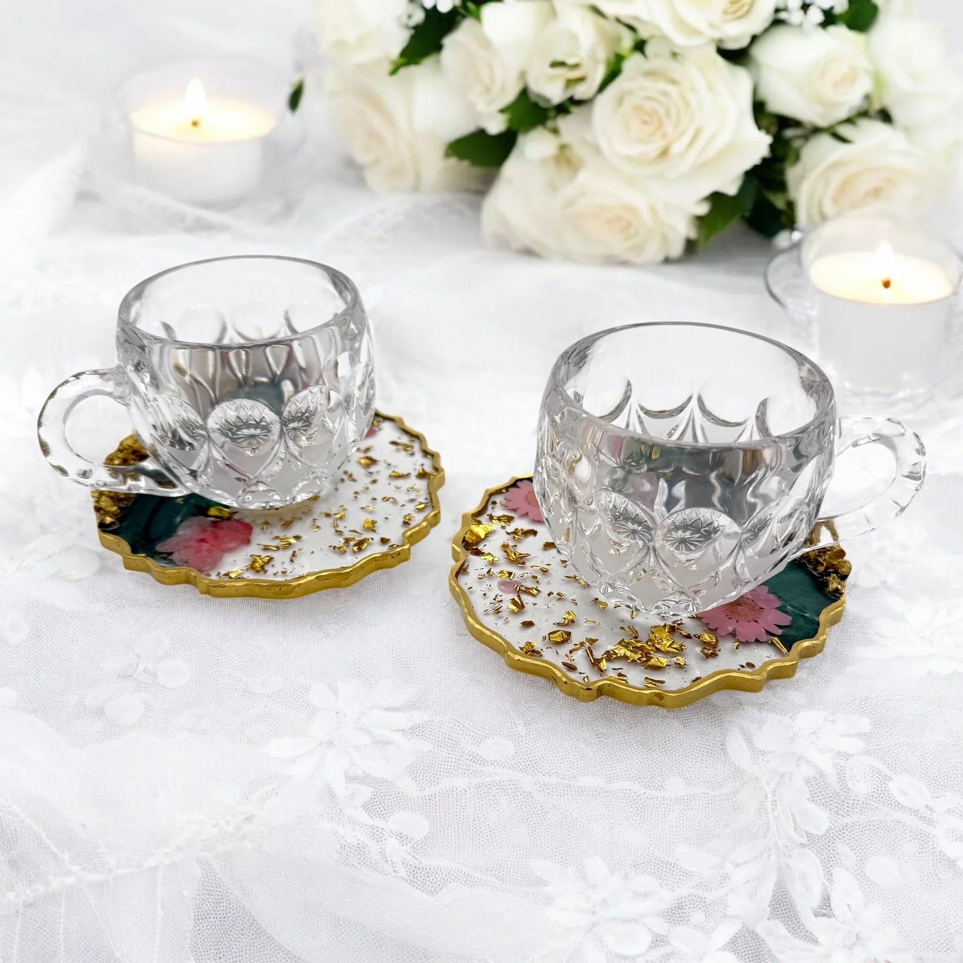 Zwei dekorative Resin Untersetzer als Unterlage für gläserne Kaffeetassen auf einer weißen Tischdecke mit Kerzen und Rosen im Hintergrund.