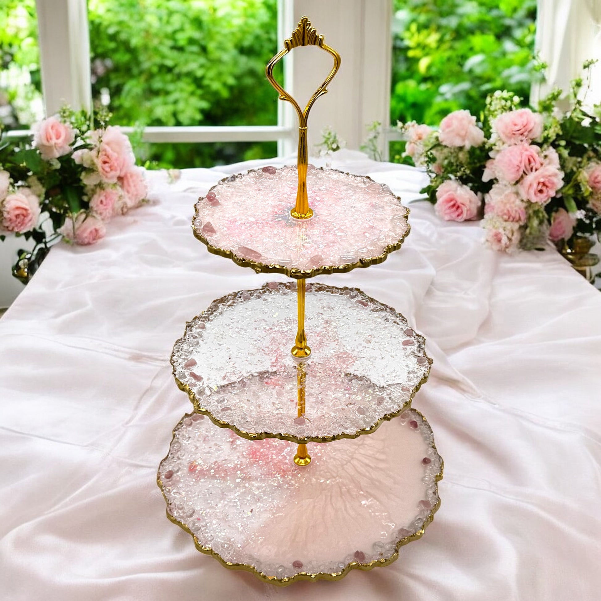 Eine dreistöckige Etagere aus rosa-transparentem Epoxidharz mit goldener Halterung und welligen Goldrändern, platziert auf einem mit weißem Stoff bedeckten Tisch, dekoriert mit rosa Rosensträußen vor einem hellen Fenster.