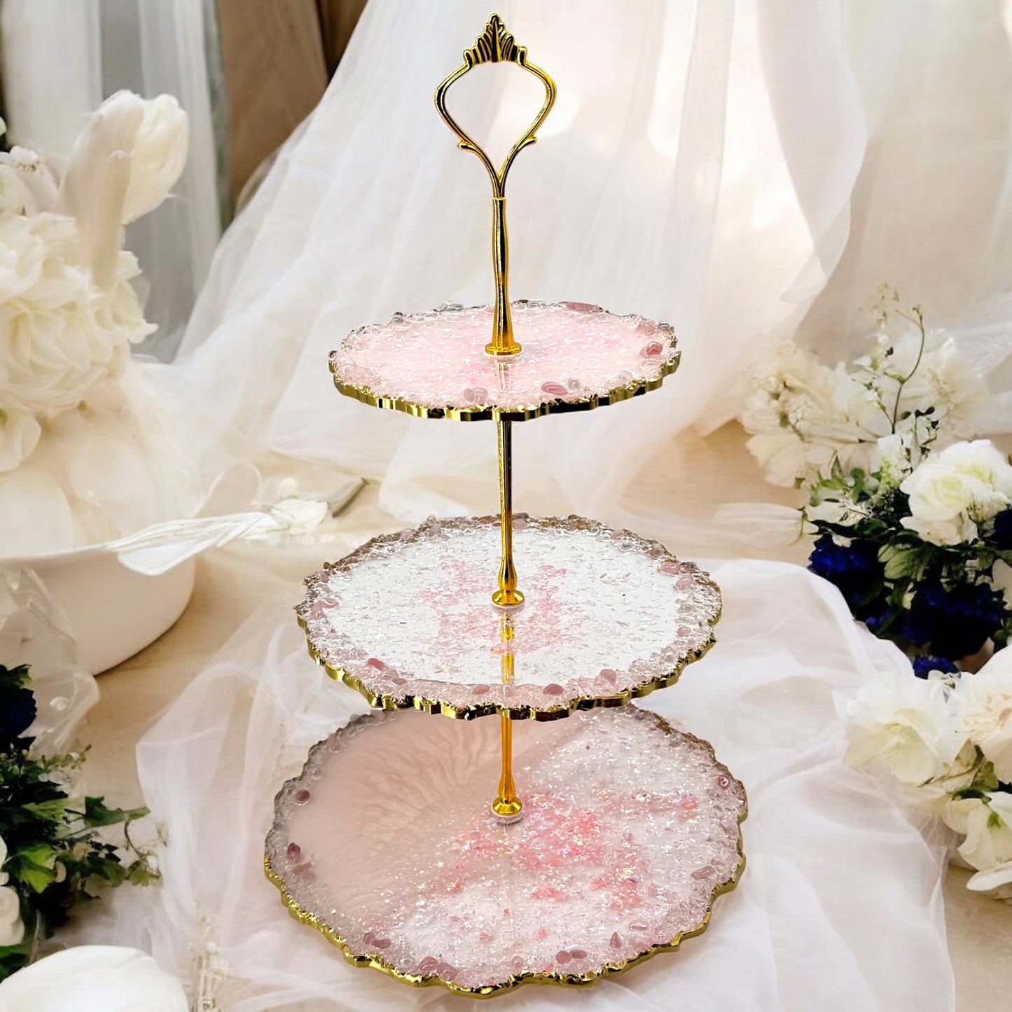 Ein dreistöckiger Etagere-Kuchenständer aus rosa und transparentem Epoxidharz mit goldener Halterung und welligen, goldverzierten Rändern, dekoriert mit kleinen rosafarbenen Steinen, platziert vor einem weißen, transparenten Stoffhintergrund mit Blumen.