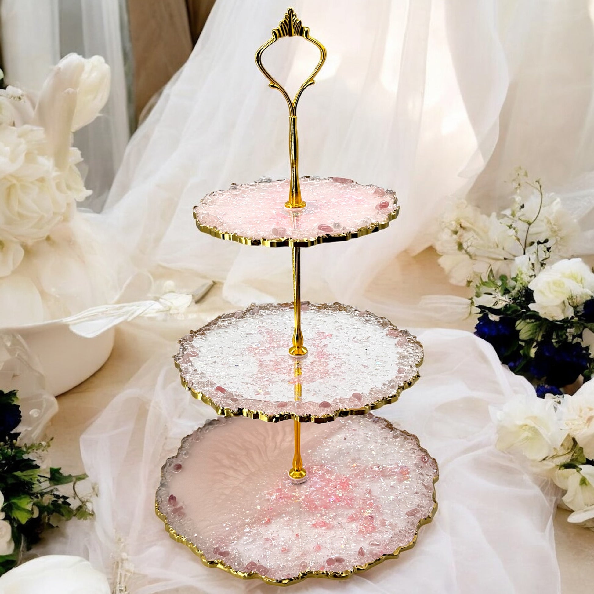 Ein dreistöckiger Etagere-Kuchenständer aus rosa und transparentem Epoxidharz mit goldener Halterung und welligen, goldverzierten Rändern, dekoriert mit kleinen rosafarbenen Steinen, platziert vor einem weißen, transparenten Stoffhintergrund mit Blumen.