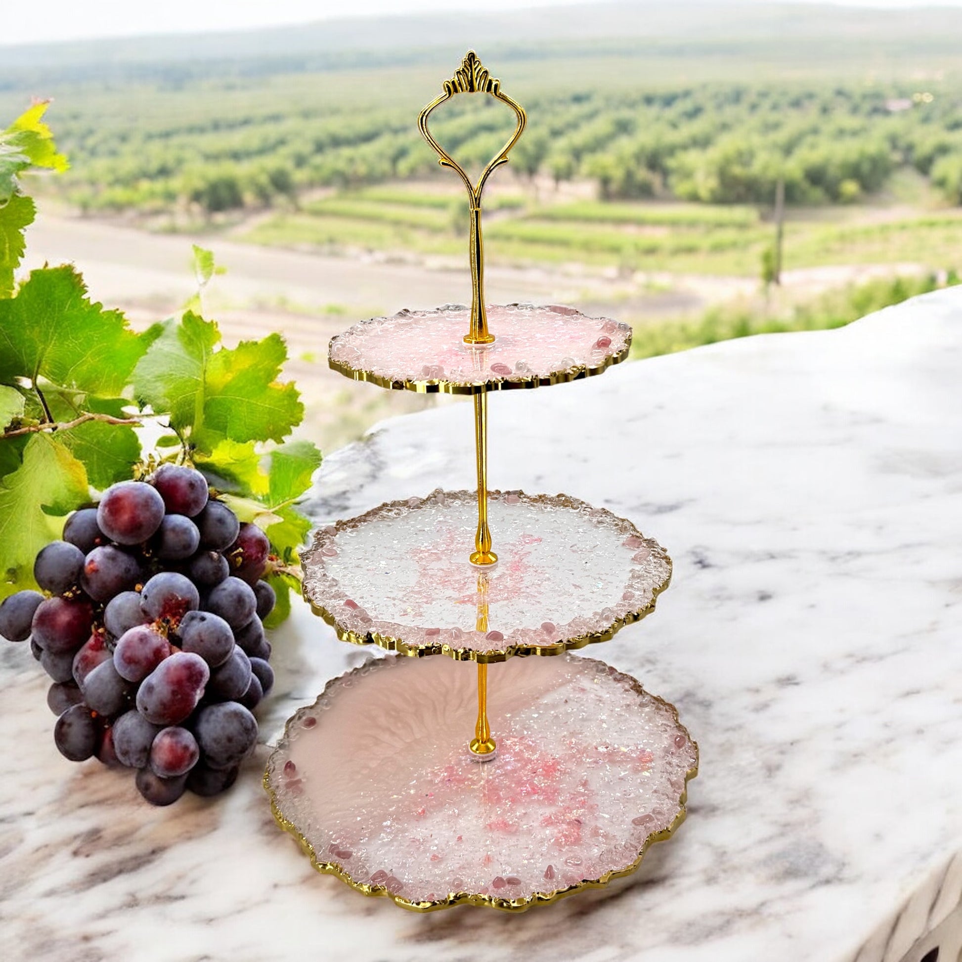 Eine dreistöckige Etagere aus rosa-transparentem Epoxidharz mit goldener Halterung und welligen Goldrändern, platziert auf einer Marmoroberfläche neben einer großen Traube dunkler Weintrauben im Freien vor einer Weinberg-Landschaft.