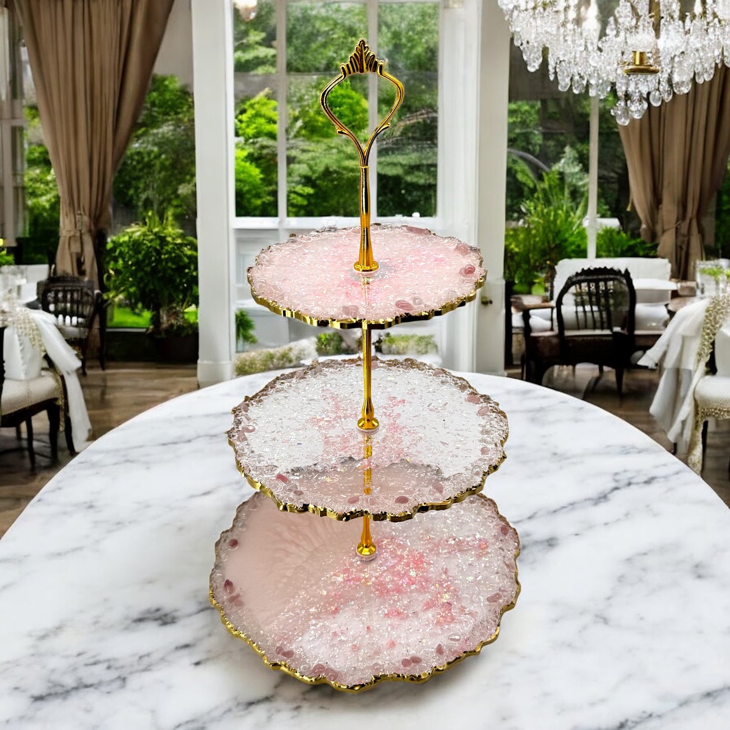 Eine dreistöckige Etagere aus rosa und transparentem Epoxidharz mit goldener Halterung und welligen Goldrändern, platziert in der Mitte eines großen ovalen Marmortisches im eleganten Innenraum eines Restaurants mit Blick auf den Garten.