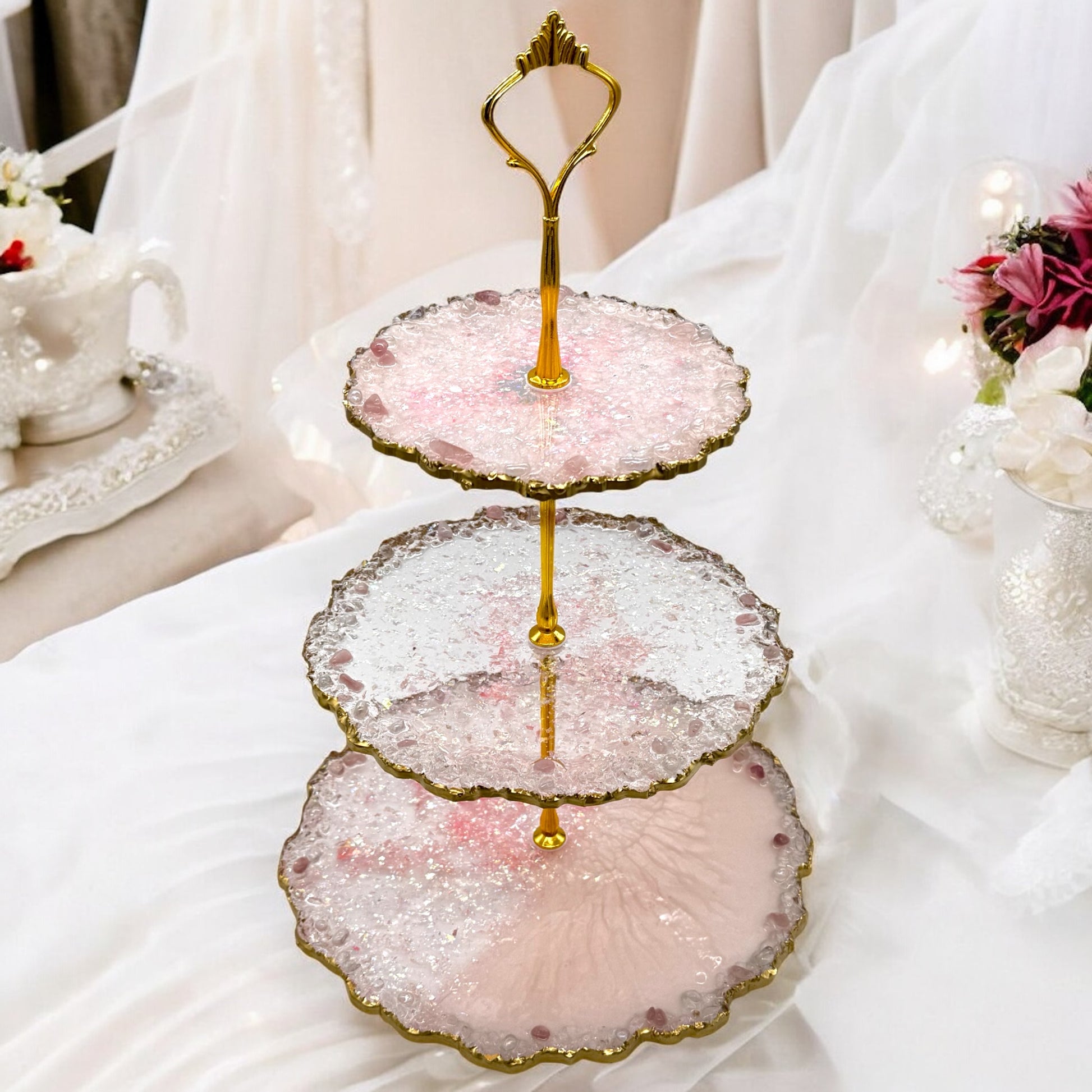 Eine dreistöckige Etagere aus rosa und transparentem Epoxidharz mit goldener Halterung, welligen Goldrändern und kleinen Dekosteinen, platziert auf einem weiß dekorierten Tisch inmitten einer festlichen Dekoration mit Blumen.