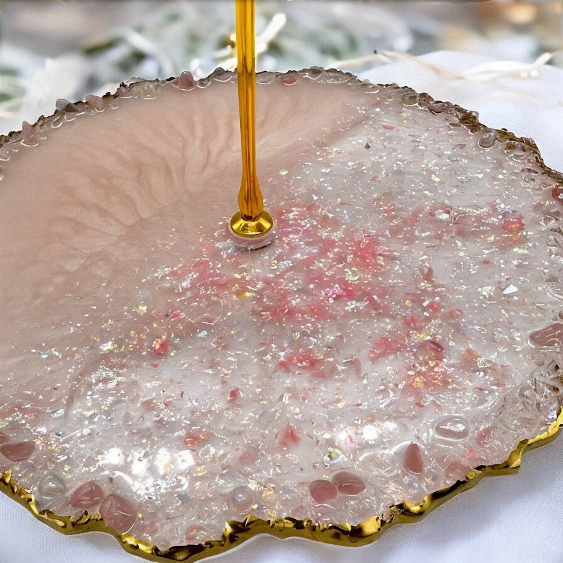 Nahaufnahme der untersten Ebene einer Etagere aus rosa Epoxidharz mit goldener Metallstange, glitzernden Elementen, unregelmäßigen Goldrändern und kleinen, am Rand eingebetteten rosafarbenen Dekosteinen auf hellem Stoffhintergrund.