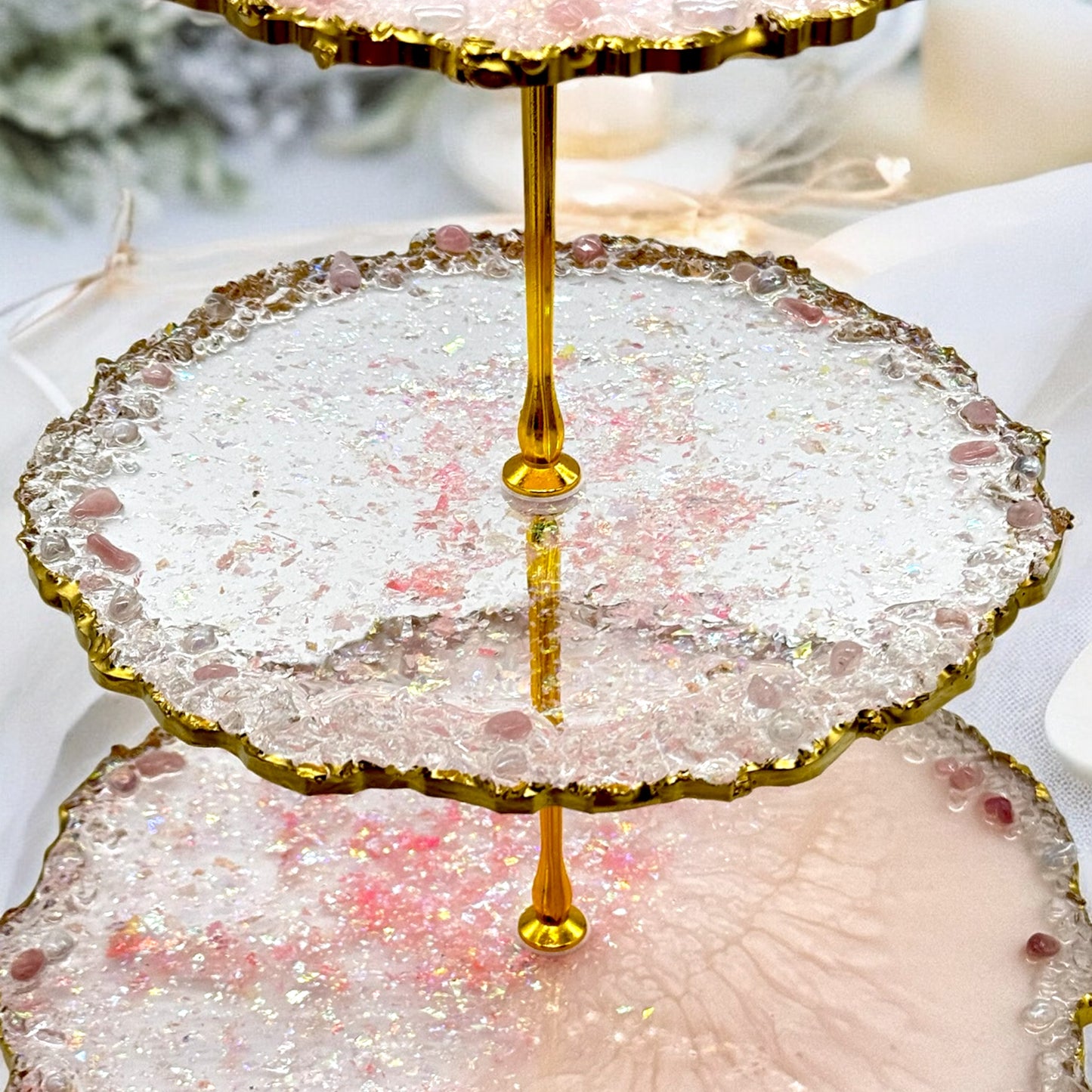 Nahaufnahme einer dreistöckigen Etagere aus klarem und rosa Epoxidharz mit goldener Metallstange, glitzernden Einschlüssen, welligen Goldrändern und kleinen eingebetteten rosafarbenen Dekosteinen auf den Etagenflächen.