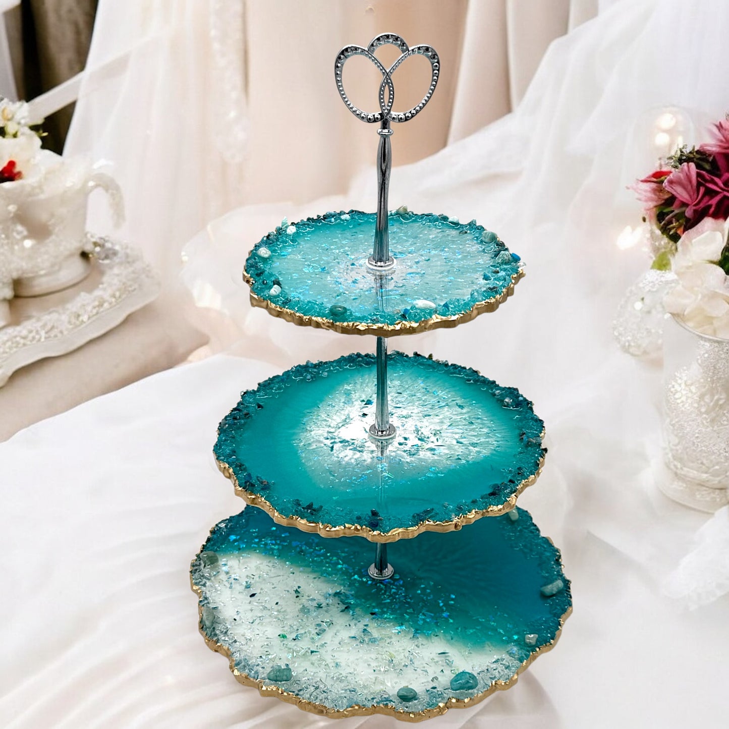 Eine dreistöckige Etagere aus türkisblauem und transparentem Epoxidharz im Geoden-Stil mit einer silbernen Metallhalterung, gewellten goldverzierten Rändern und eingebetteten Dekosteinen, platziert auf einem weißen, eleganten Stoffhintergrund inmitten von Hochzeitsdekorationen.