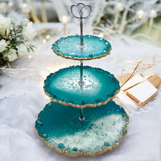 Eine dreistöckige Etagere aus türkisblauem und transparentem Epoxidharz im Geoden-Stil mit einer silbernen Metallhalterung, gewellten goldverzierten Rändern und eingebetteten Dekosteinen, platziert auf einem weißen, strukturierten Stoffhintergrund.
