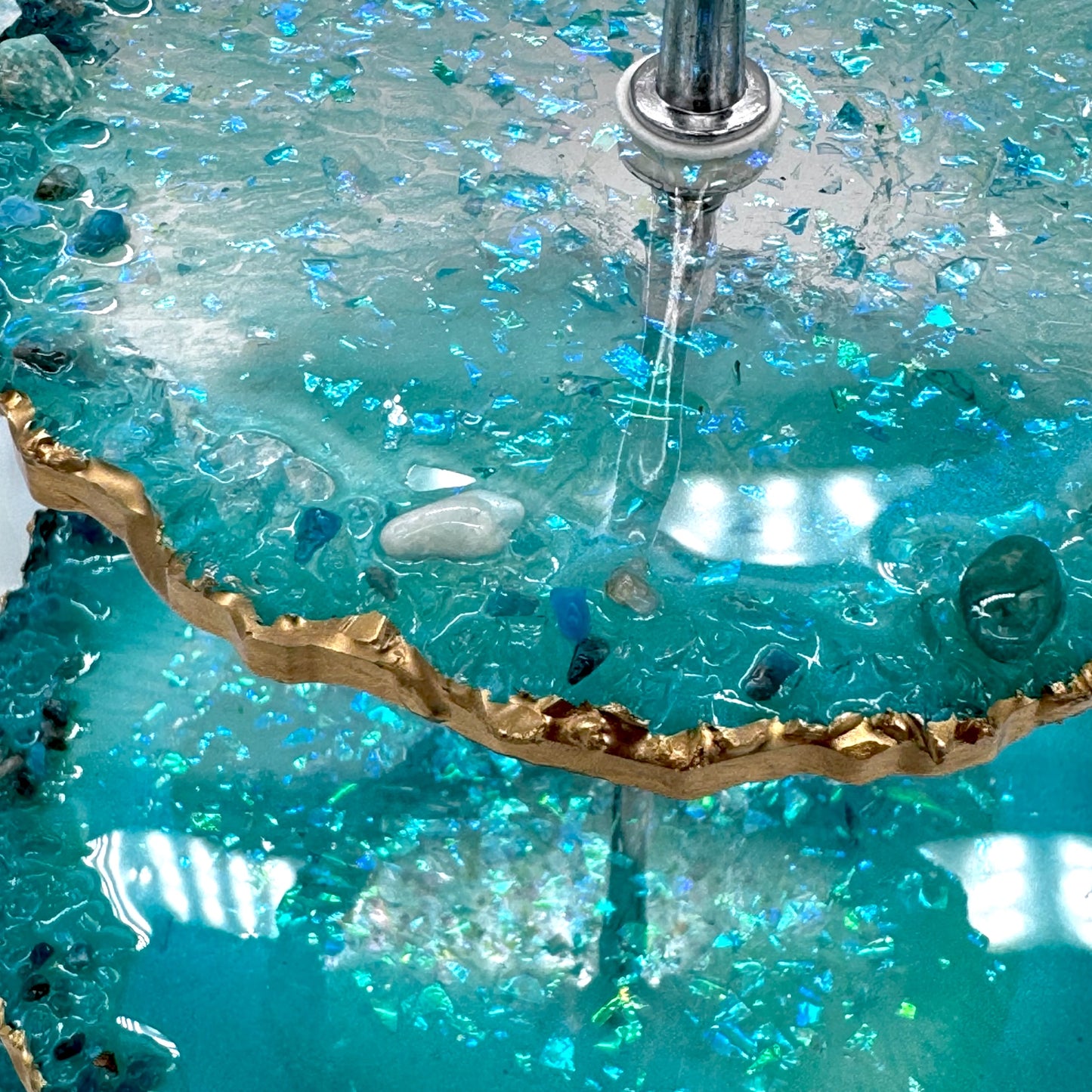 Nahaufnahme einer Etagere aus türkisblauem Epoxidharz im Geoden-Stil mit einer silbernen Metallstange, glitzernden blauen Foliensplittern, unregelmäßigen goldverzierten Rändern und eingebetteten kleinen Dekosteinen auf den Etagenflächen.