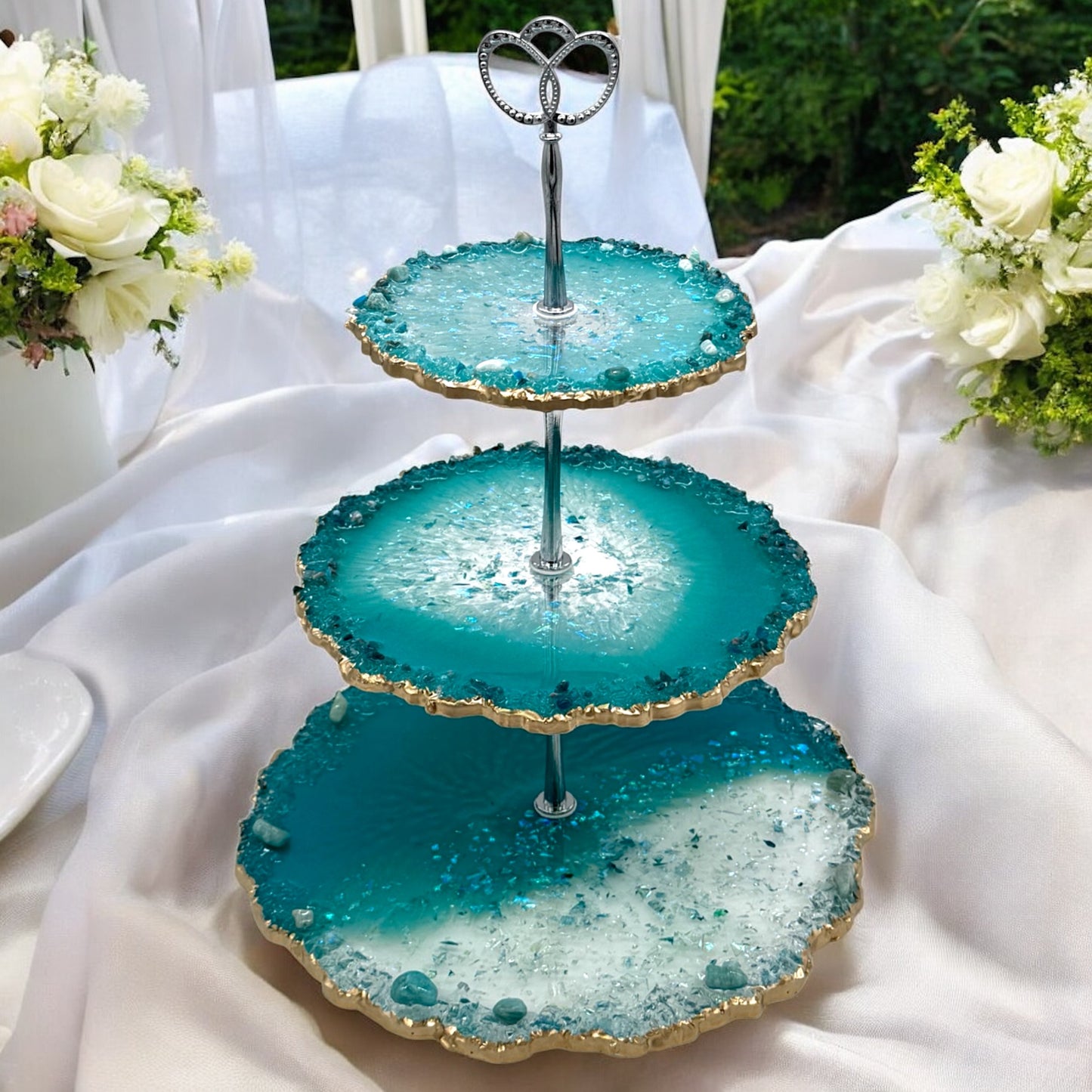 Eine dreistöckige Etagere aus türkisblauem und transparentem Epoxidharz im Geoden-Stil mit einer silbernen Metallhalterung und gewellten goldverzierten Rändern, platziert auf einem weißen Stofftisch im Freien, flankiert von zwei weißen Rosensträußen