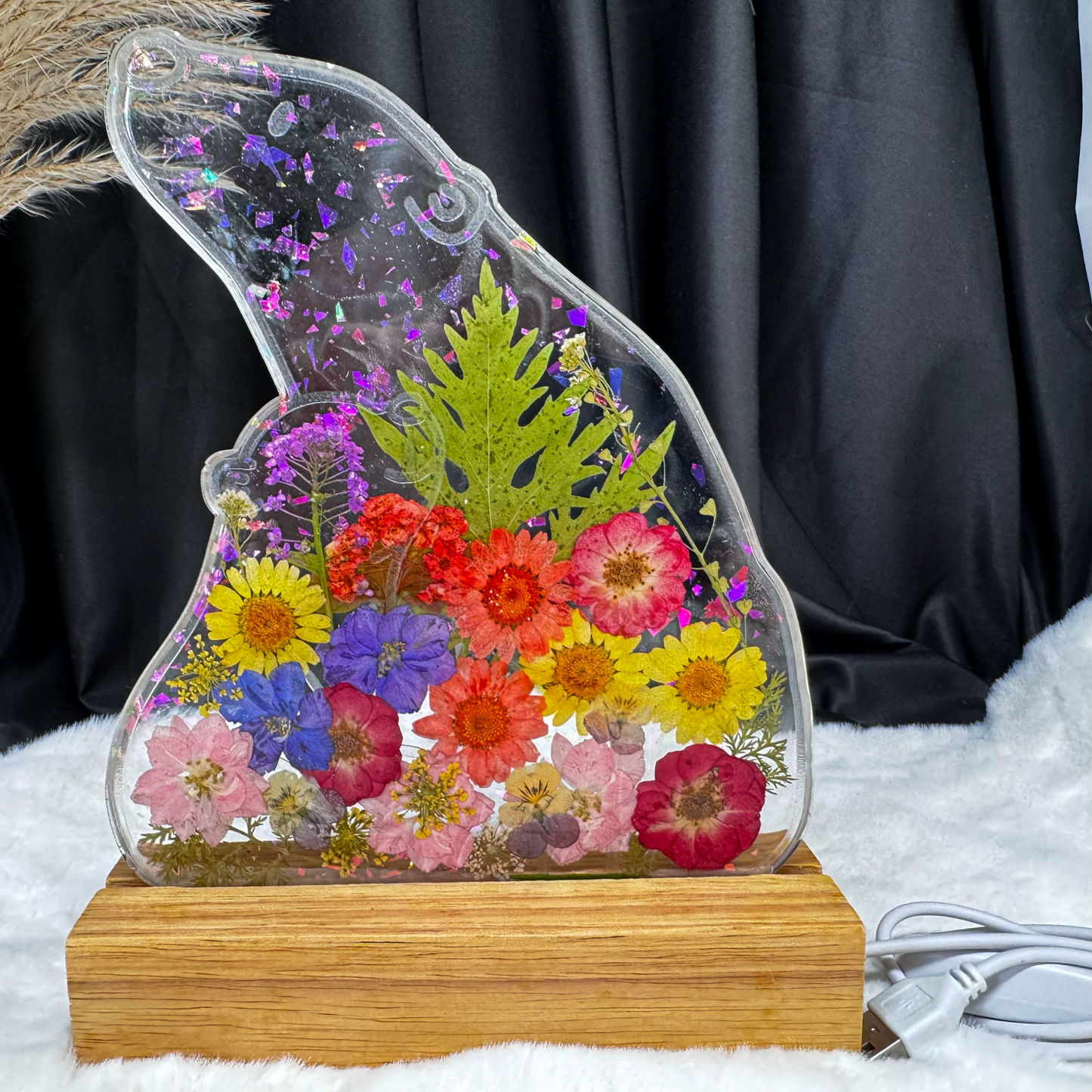 Ein bärenförmiges Epoxidharz-Nachtlicht, reich gefüllt mit bunten Trockenblumen und glitzernden lila Partikeln im oberen Bereich, steht ausgeschaltet auf einem Holzsockel vor einem dunklen Vorhang.