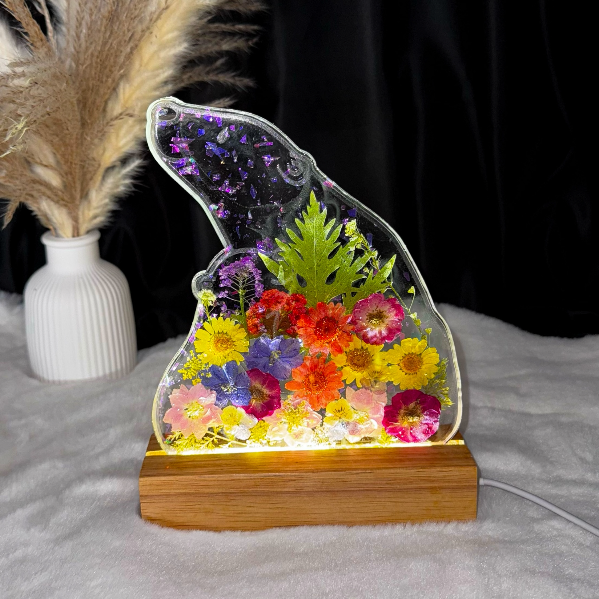 Ein bärenförmiges Epoxidharz-Nachtlicht mit bunten Trockenblumen leuchtet auf einem Holzsockel vor einem dunklen Hintergrund, während im Hintergrund eine weiße Vase mit Pampasgras auf einer weichen, weißen Decke steht.