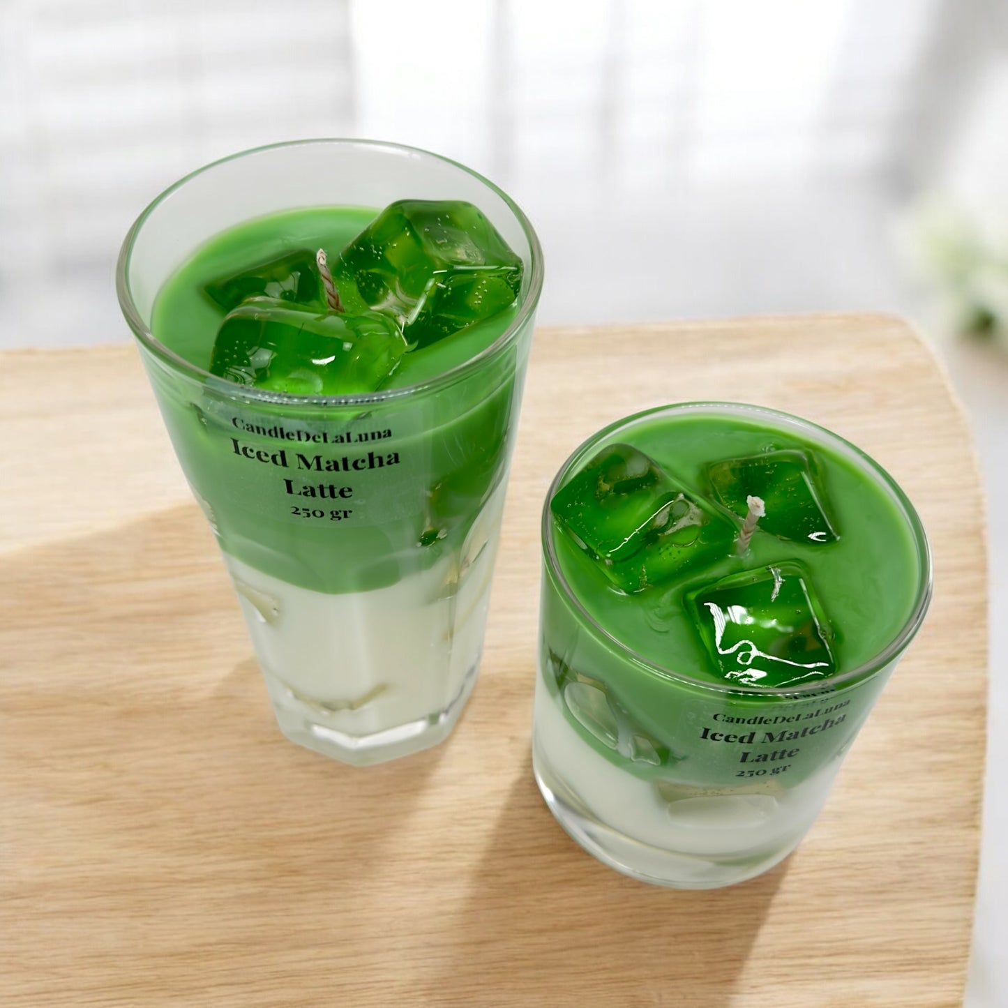 Zwei handgemachte Kerzen im Design von Iced Matcha Latte in einem hohen und einem niedrigen Glas mit grün-weißen Wachsschichten, künstlichen grünen Eiswürfeln auf der Oberseite und der Aufschrift „CandleDeLuna Iced Matcha Latte 250 gr“, platziert auf einer Holzoberfläche vor einem hellen Hintergrund.