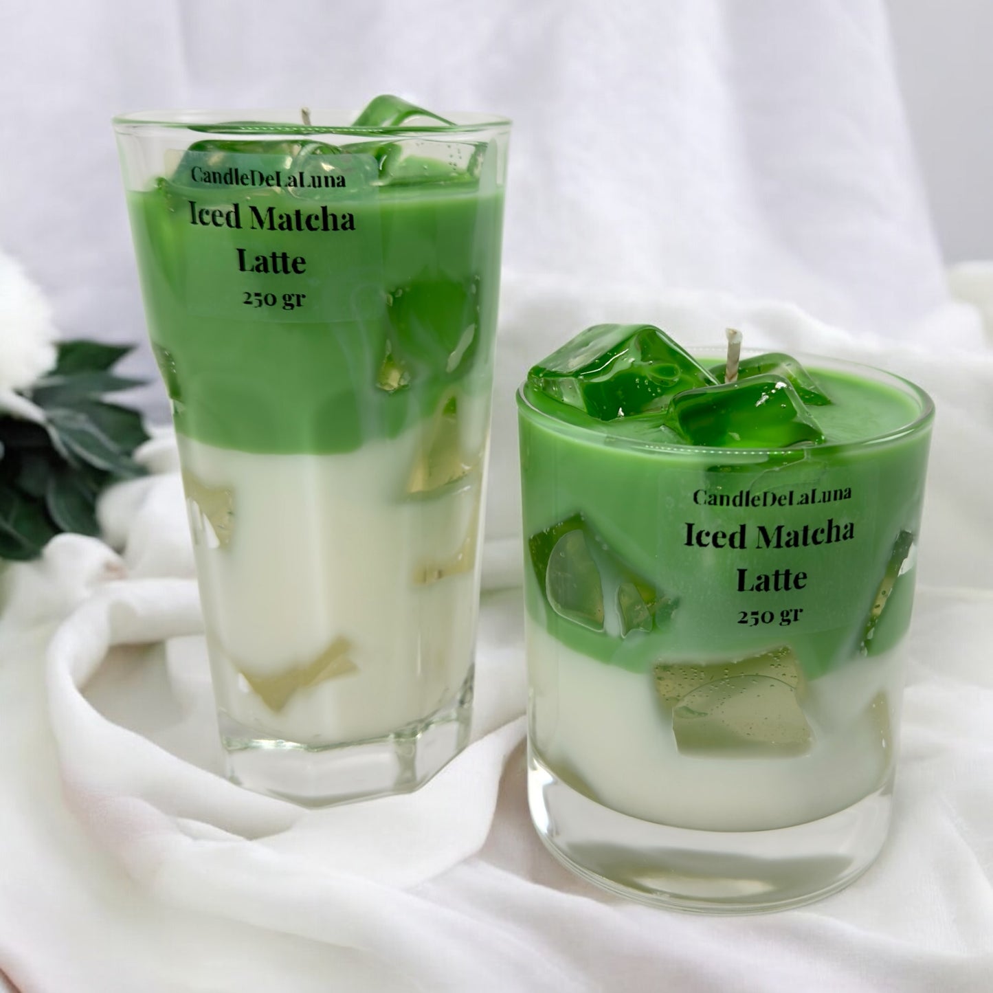 Zwei handgemachte Kerzen im Design von Iced Matcha Latte in einem hohen und einem niedrigen Glas mit grün-weißen Wachsschichten, künstlichen Eiswürfeln aus Wachs und der Aufschrift „CandleDeLuna Iced Matcha Latte 250 gr“ auf einem weißen Stoffhintergrund.