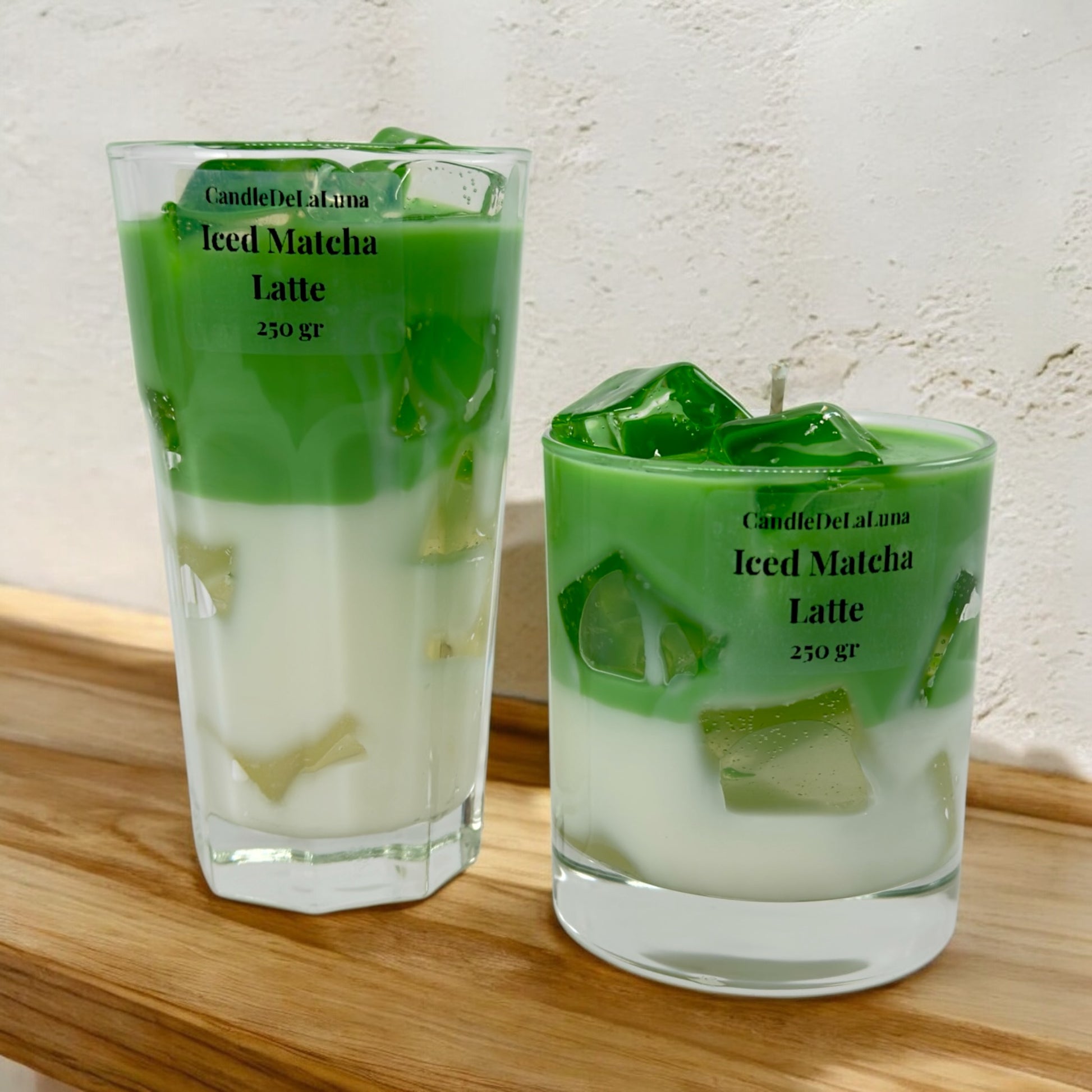 Zwei handgemachte Kerzen im Design von Iced Matcha Latte, präsentiert in einem hohen facettierten Glas und einem niedrigen runden Glas mit grün-weißen Wachsschichten und künstlichen Eiswürfeln auf einer Holzoberfläche vor einer strukturierten weißen Wand.