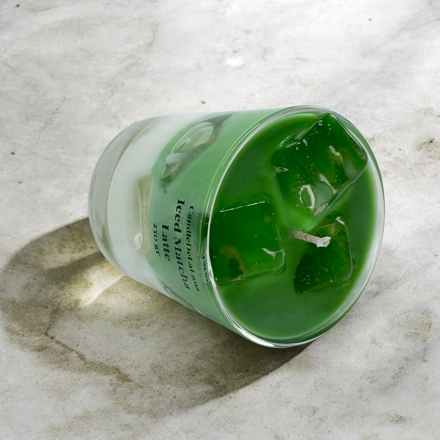 Eine handgemachte Kerze im Design von Iced Matcha Latte in einem niedrigen Glas, gefüllt mit grünem und weißem Wachs sowie künstlichen grünen Eiswürfeln auf der Oberseite, liegend auf einer hellen Marmoroberfläche mit Schattenwurf.